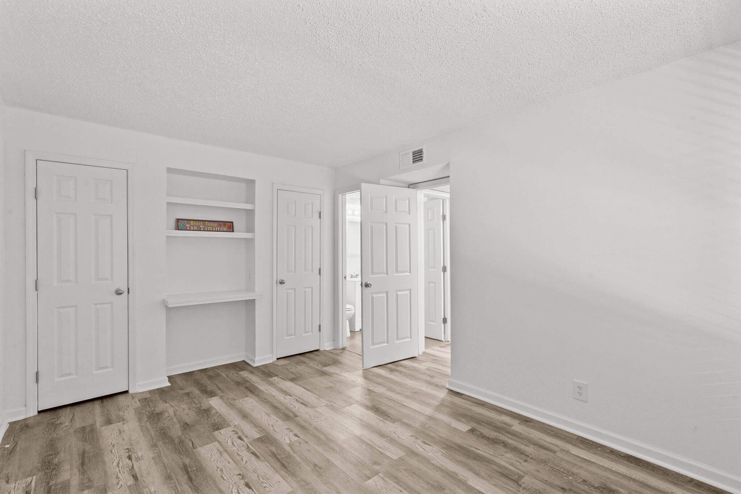 A bright, empty room featuring light-colored walls, wooden flooring, and two closed doors on the left. There’s a small built-in shelf on the wall. A third open door leads to a bathroom area, providing a spacious and clean look. Natural light streams in, creating an inviting atmosphere.