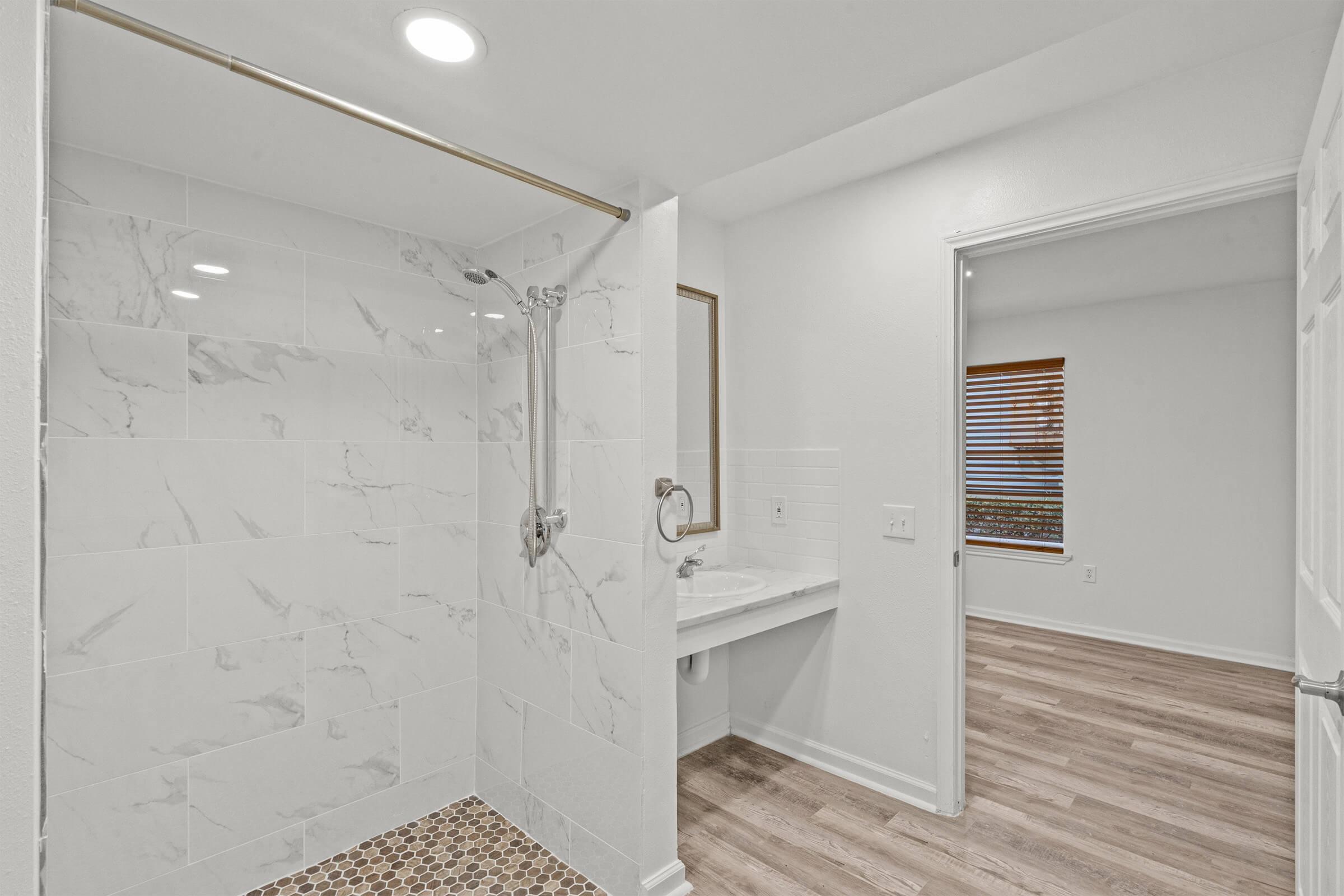 Modern bathroom featuring a glass-enclosed shower with marble tiles, a stylish showerhead, and a mosaic floor. There is a vanity area with a mirror and a doorway leading to another room with window blinds and wooden flooring. The walls are painted in a light color, creating a bright and airy feel.