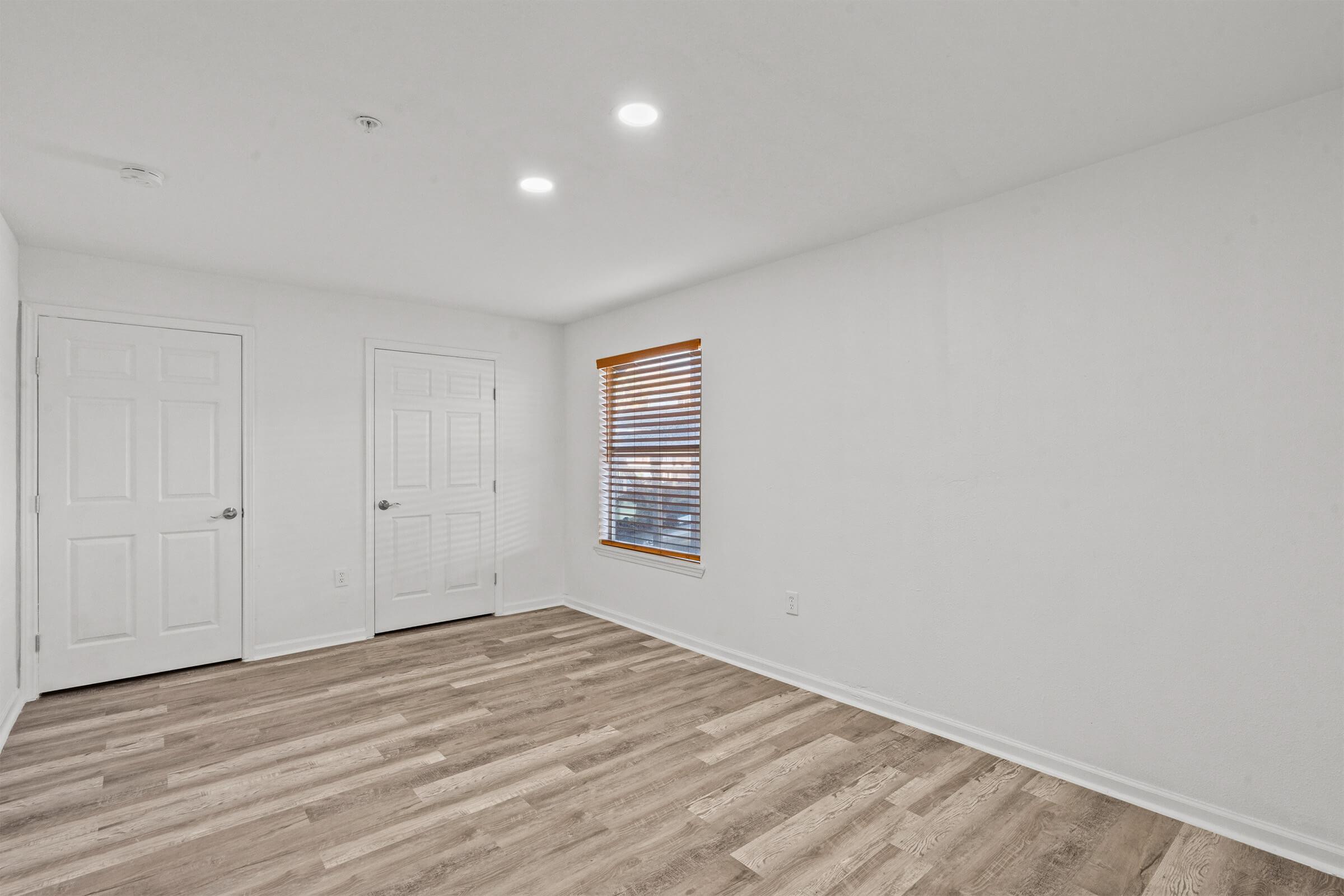 A minimalist room featuring light-colored walls and wooden flooring. There are two closed doors on the left and a window with blinds on the right, allowing natural light to enter. The ceiling has recessed lighting, creating a bright ambiance in the space.