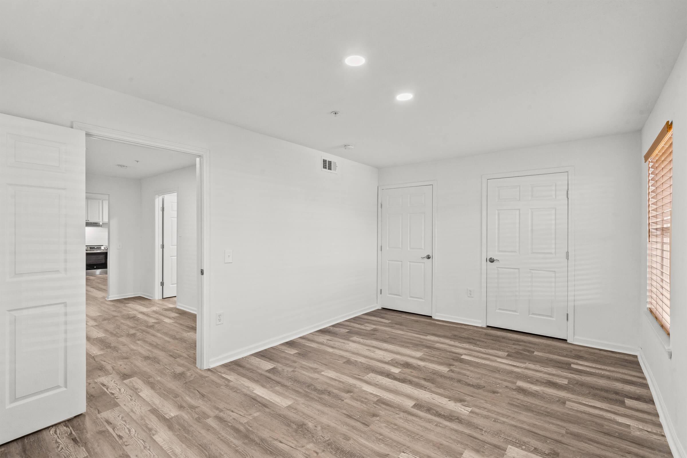 A spacious, unfurnished room with light-colored walls and hardwood-like flooring. Two doors are visible, one leading to another room and the other possibly to a closet. There are recessed ceiling lights illuminating the space, and a window with blinds allows natural light to enter.