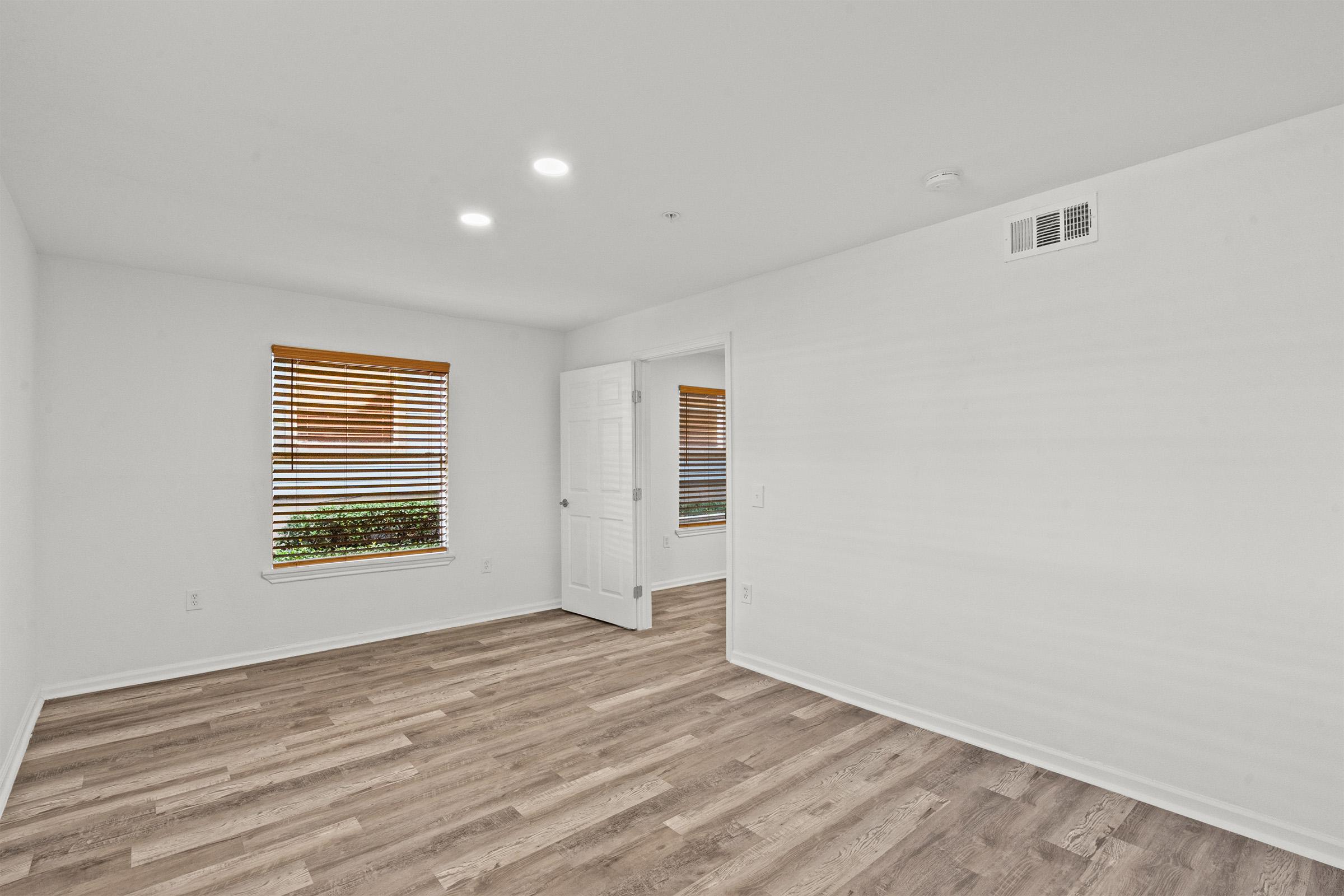 A bright, empty room featuring light-colored walls, wooden flooring, and a window with blinds. The room has recessed lighting and an open door leading to another space, creating a sense of spaciousness and minimalism.