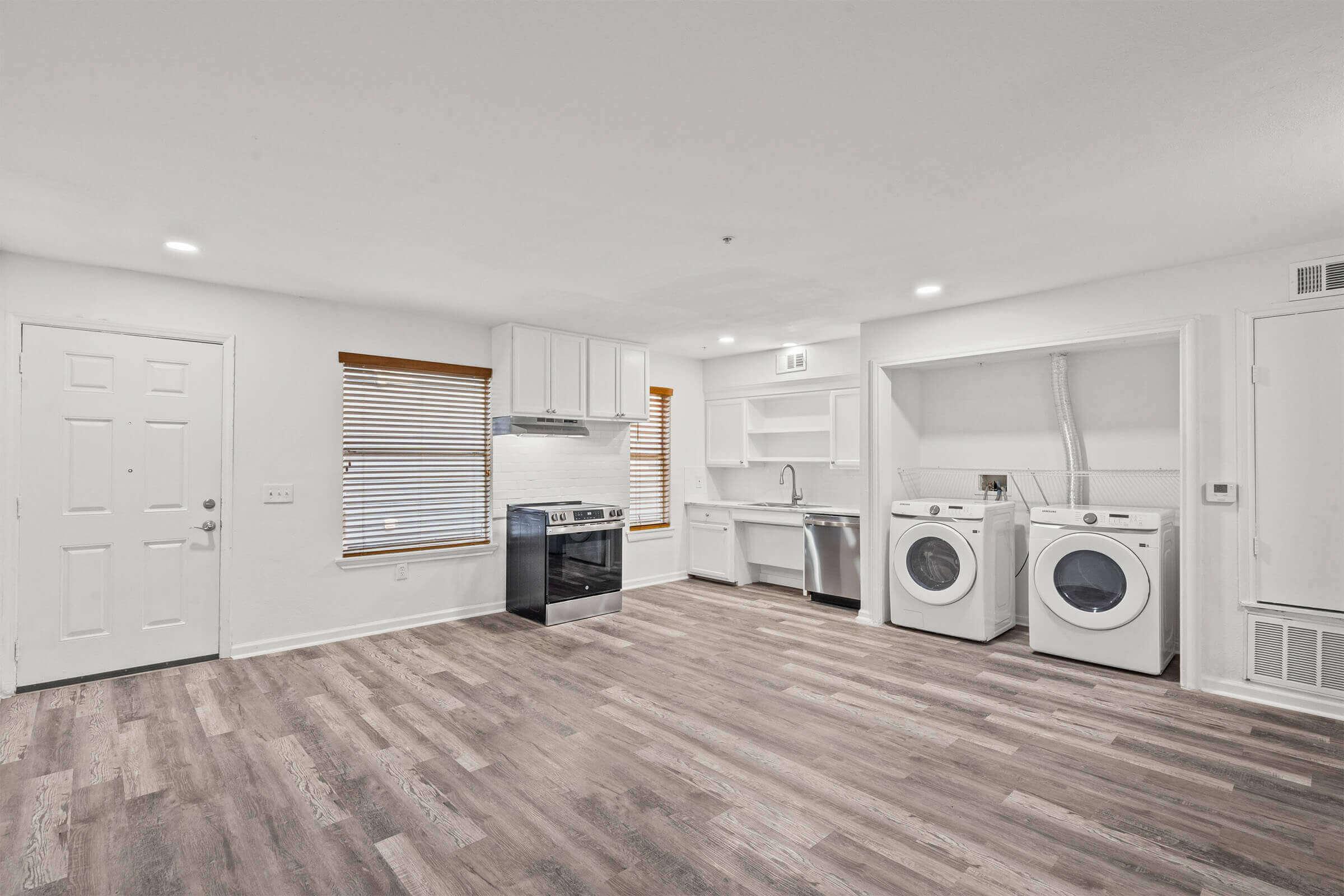 A spacious, modern kitchen and laundry area featuring a stainless steel oven, a sink, and white cabinetry. Two front-loading washing machines are situated next to the sink. The room has light-colored walls, wooden floorboards, and natural light coming from a window with blinds.