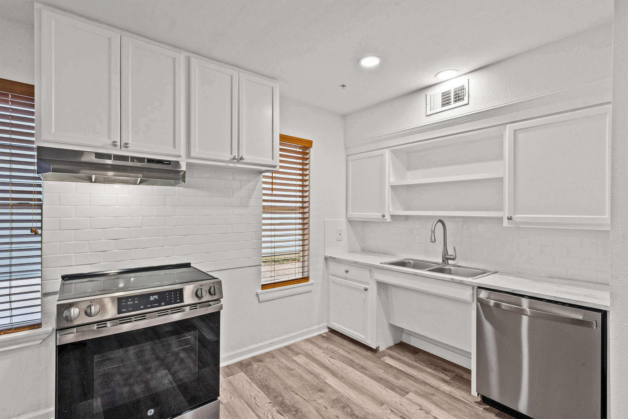 Modern kitchen featuring white cabinetry, stainless steel appliances, and a sleek tile backsplash. Natural light streams in through wooden blinds on the windows, and the space has a clean, minimalist design with a double sink and an oven range.