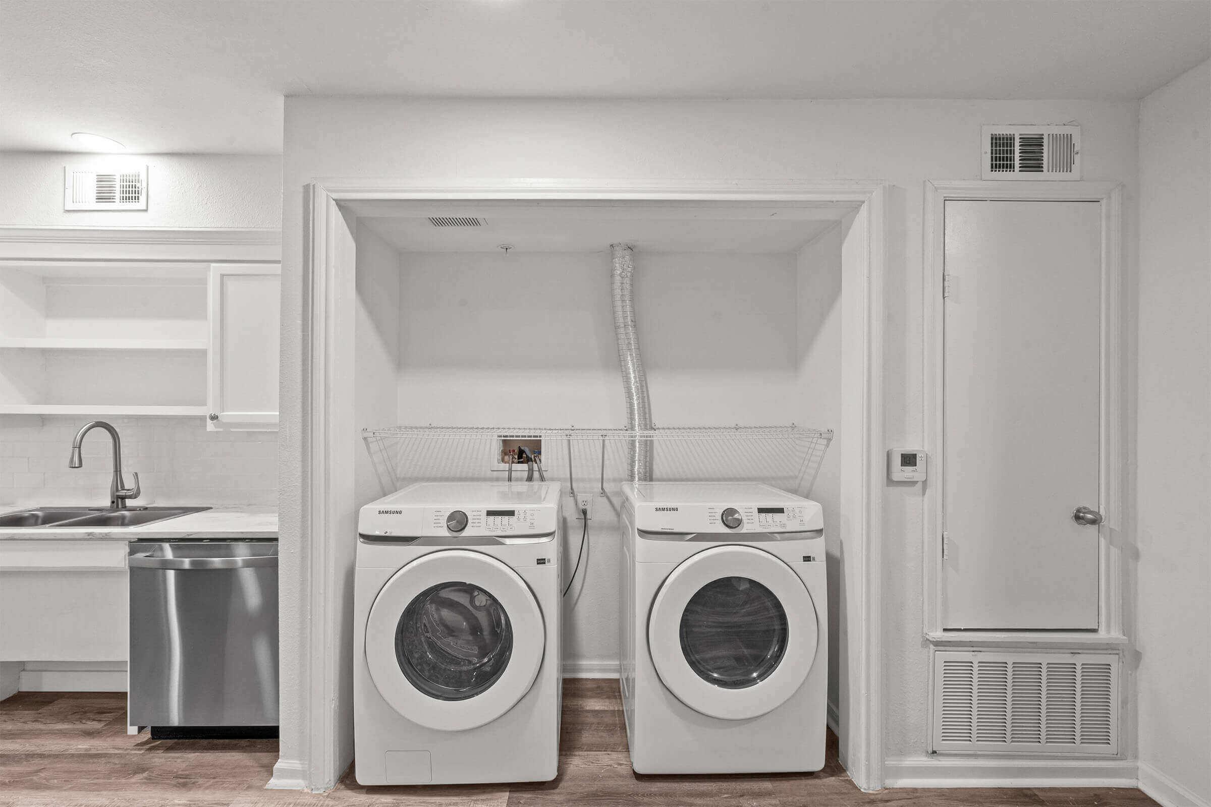 A laundry room featuring a stackable washer and dryer, both in white. The appliances are positioned in a built-in space, beside a modern kitchen sink and dishwasher. The walls are painted white, and there is a door on the right leading to another room. The floor is a warm, light wood.