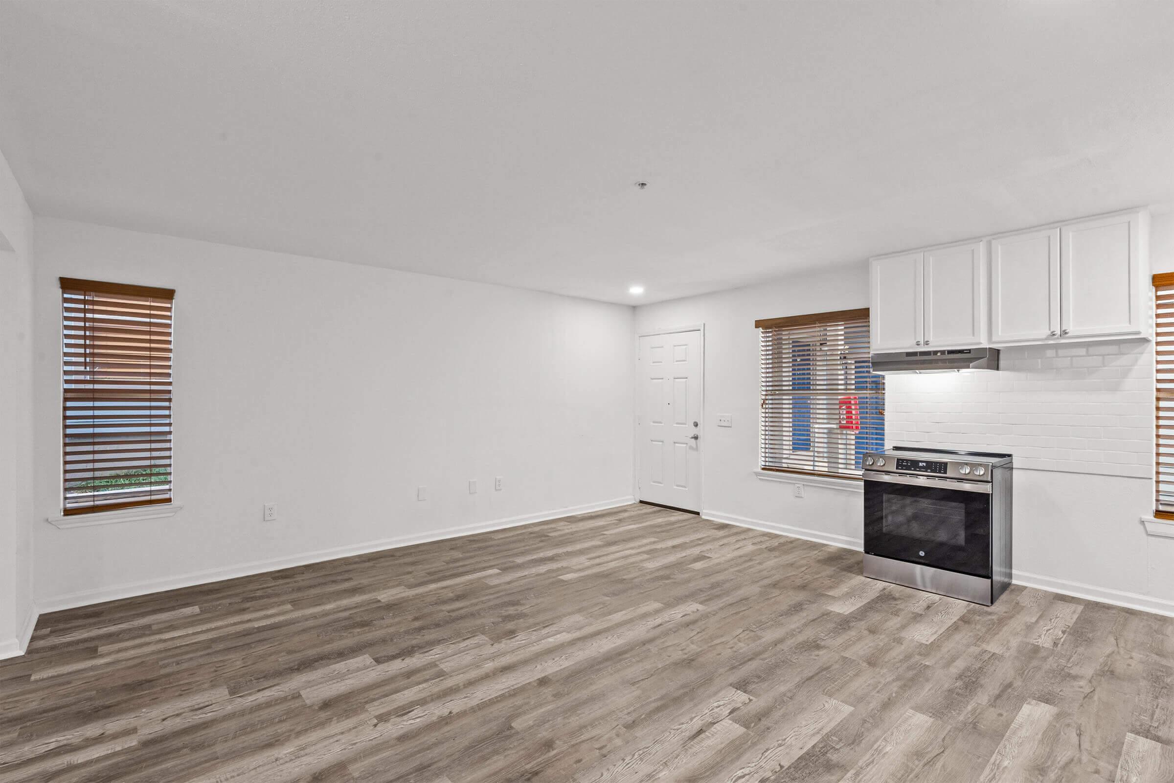 An empty, modern living space with light-colored walls and hardwood flooring. The room features two windows with wooden blinds, a white door, and a corner kitchen area with a black stove, white cabinets, and a minimalist design. Natural light illuminates the space, giving it a bright and airy feel.