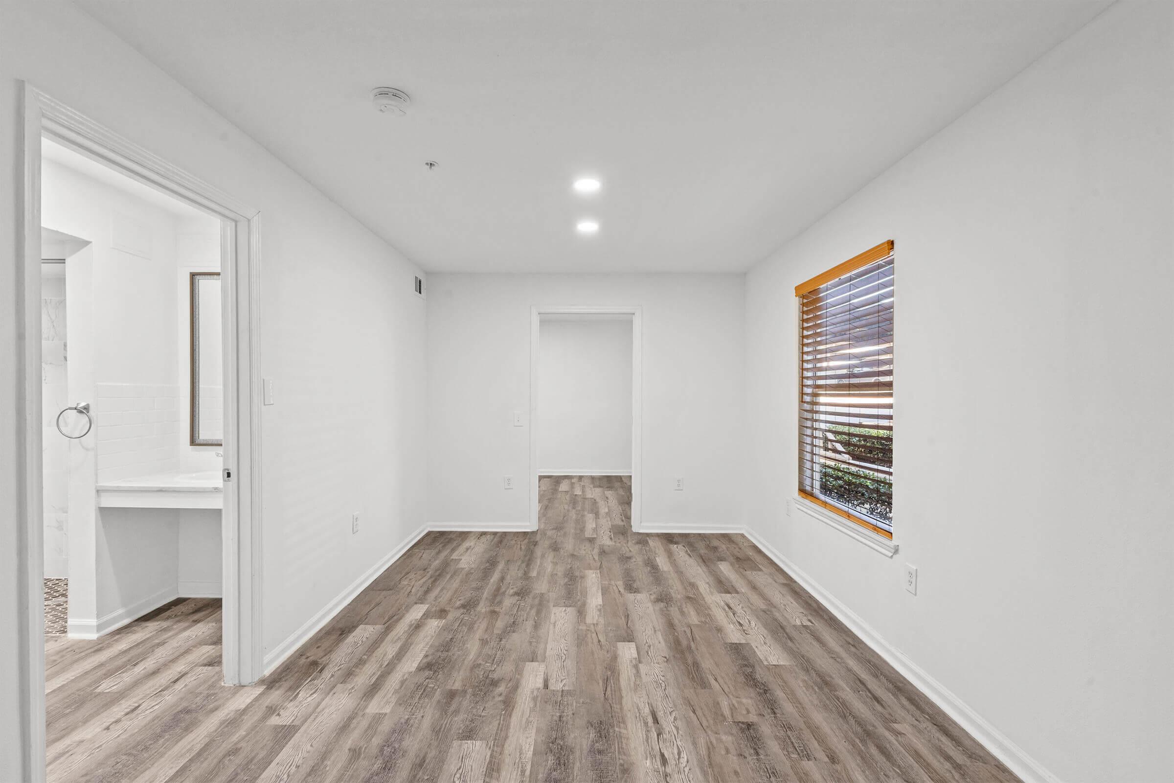 A modern, empty room with light hardwood flooring and white walls. It features a window with wooden blinds allowing natural light in, and a door that leads to a bathroom on the left. The room has recessed lighting and a spacious layout, making it suitable for various design options.