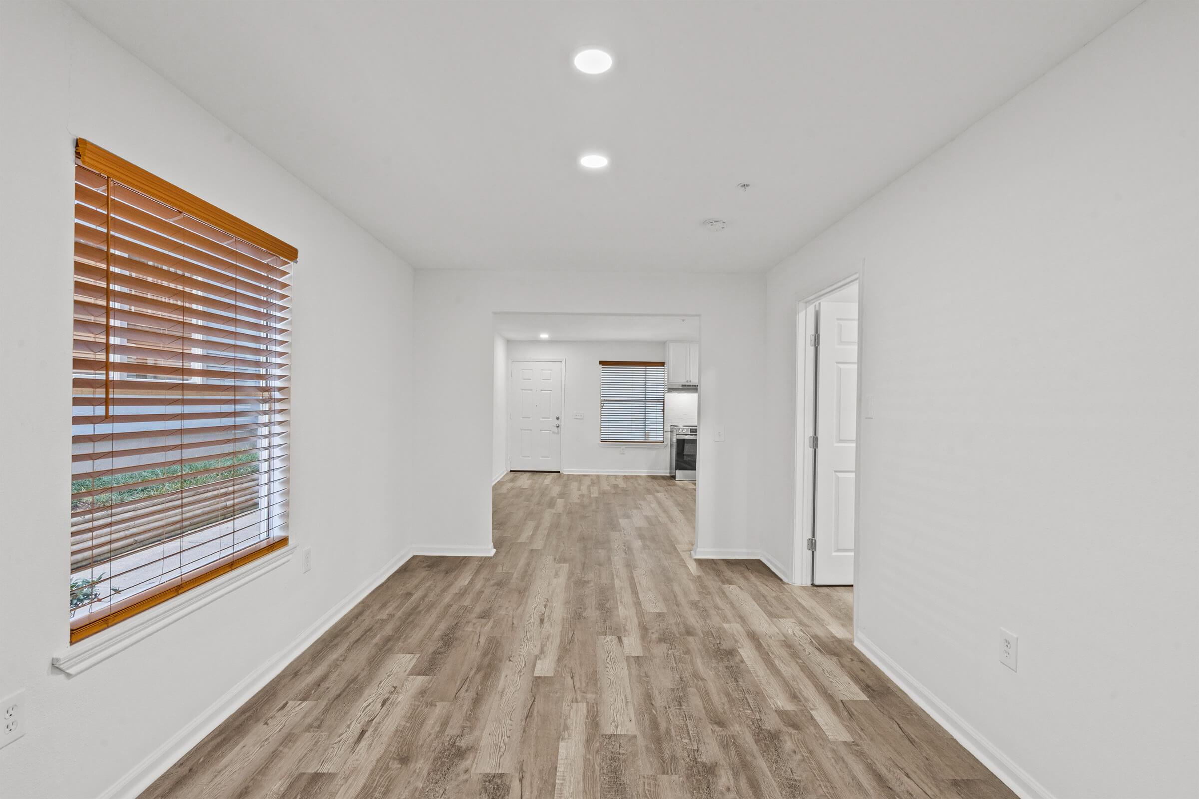 A spacious, empty room featuring light-colored walls and wooden flooring. Natural light streams in through a window with wooden blinds. A doorway leads to a kitchen area in the background, and another doorway opens to an adjacent space. There are ceiling lights providing illumination.