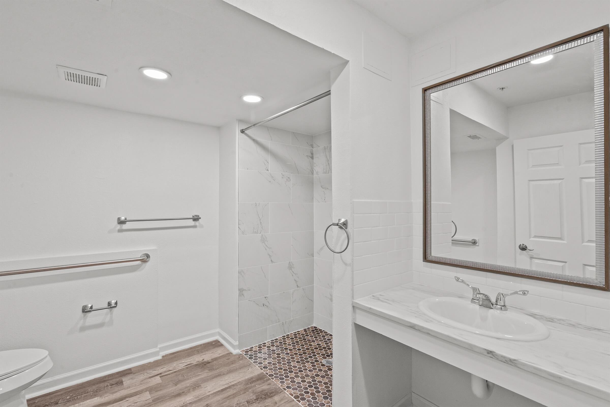 A modern bathroom featuring a white tiled shower, a mirror above a marble countertop, and a toilet. The walls are painted white, with recessed lighting and a textured floor. Grab bars are installed near the shower and toilet for accessibility.