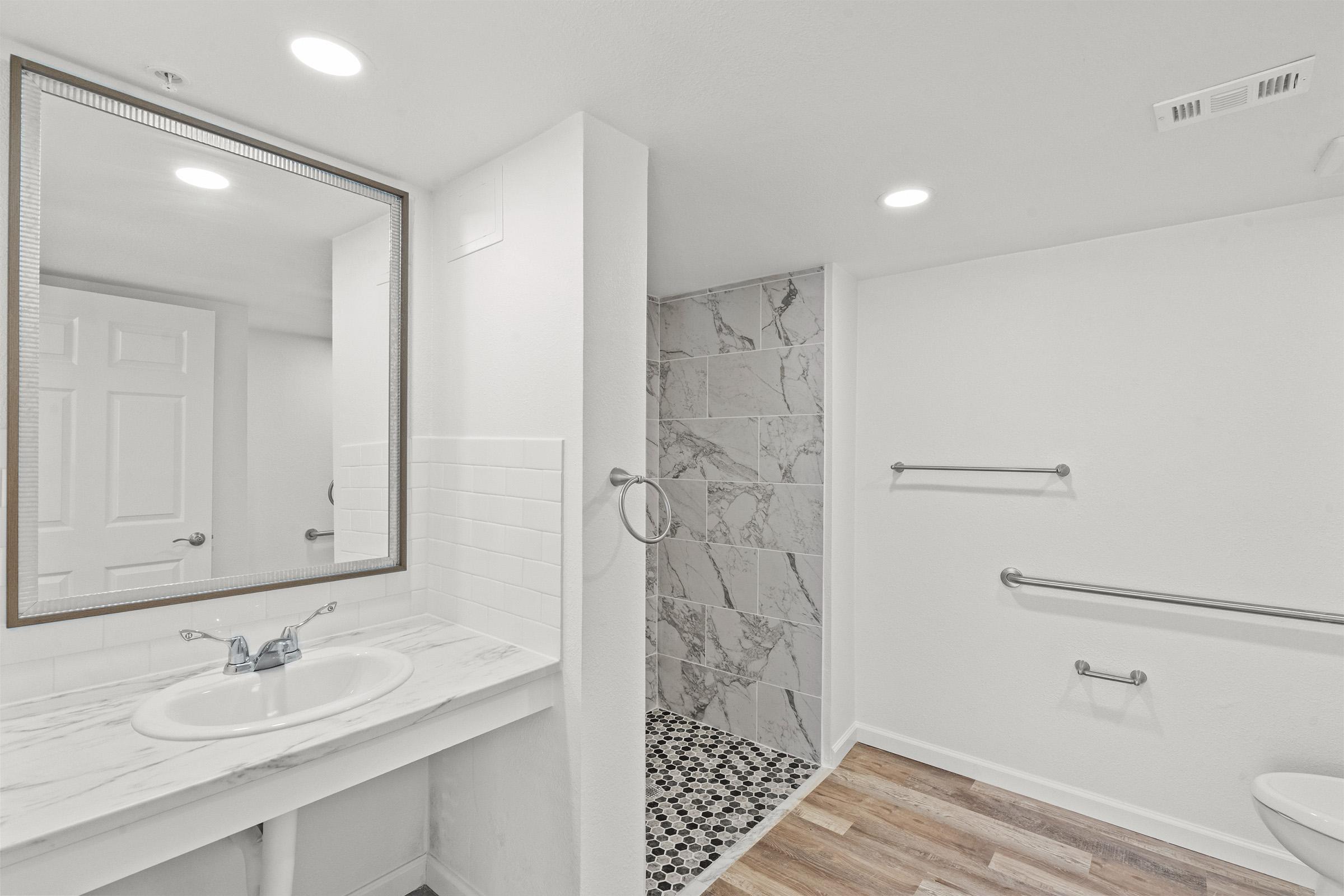 A modern restroom featuring a marble countertop sink with a mirror above, a tiled shower area with marble and patterned tiles, and wooden flooring. The space is well-lit with recessed lighting and includes grab bars for accessibility. The walls are painted in a light color, enhancing the bright and spacious feel.