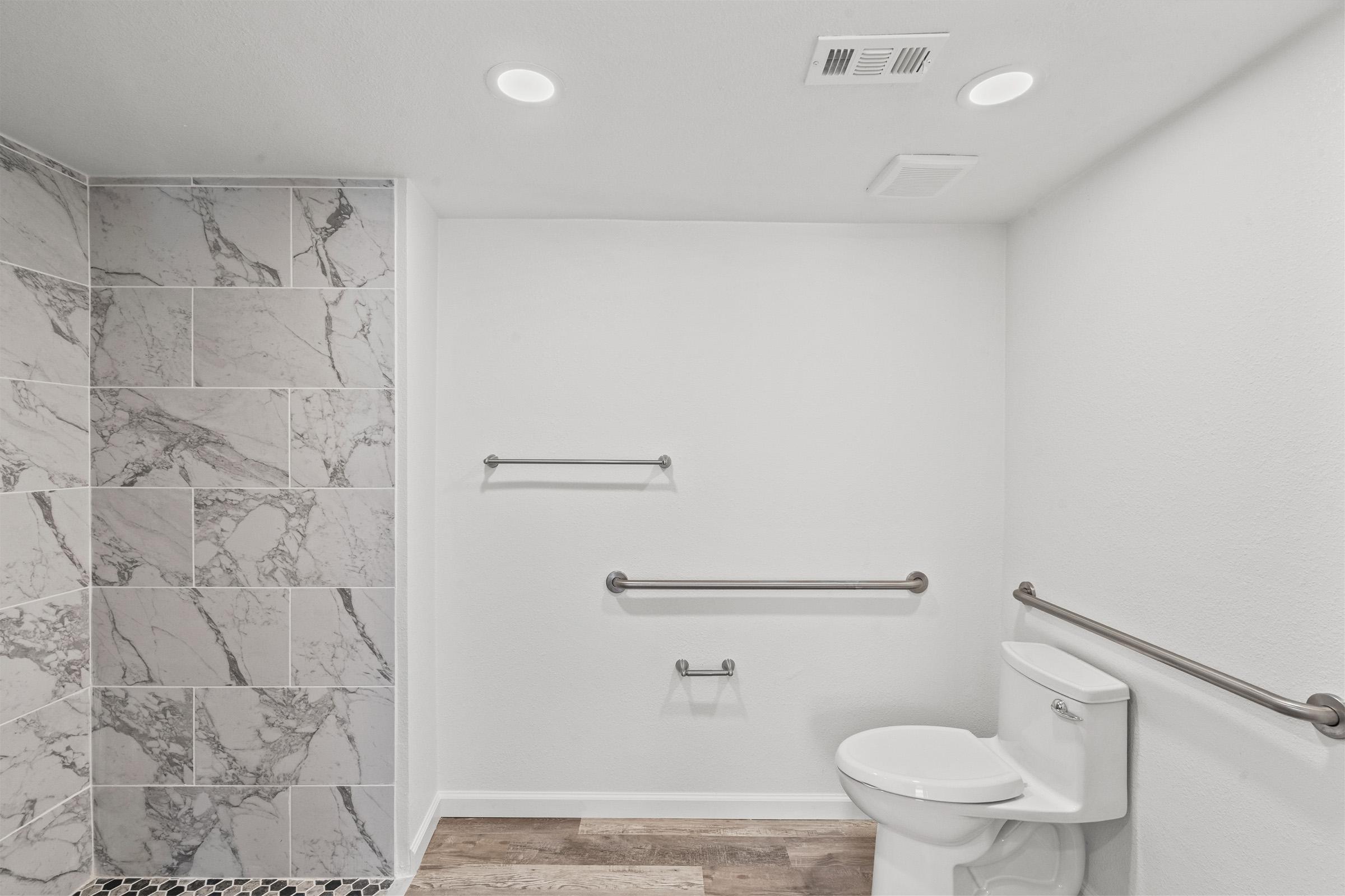 A clean, modern bathroom featuring a white toilet beside grab bars for accessibility. The wall is adorned with large marble tiles, and the floor has dark, textured tiles. The space is well-lit with recessed ceiling lights, offering a bright and spacious feel.