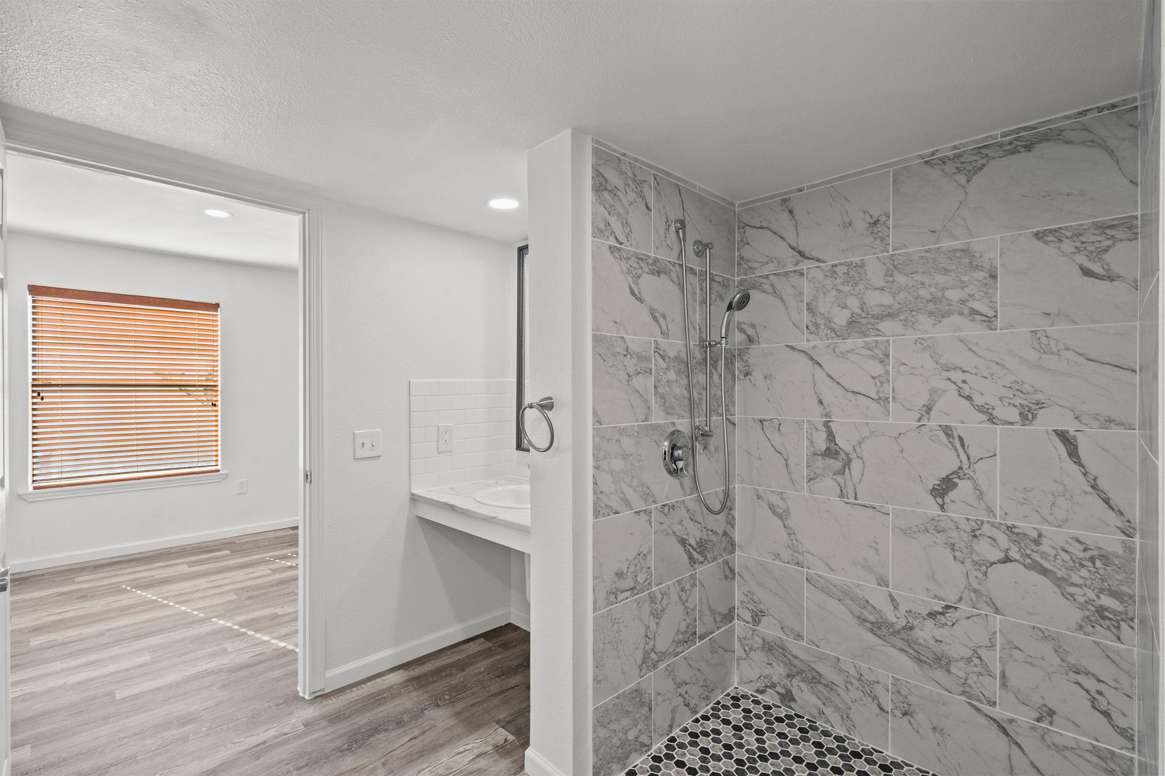 Renovated bathroom featuring a modern shower area with marble tiles, a window with wooden blinds, and a spacious view of a well-lit room beyond. The floor is adorned with wooden planks, and there is a sink area visible in the adjacent space.