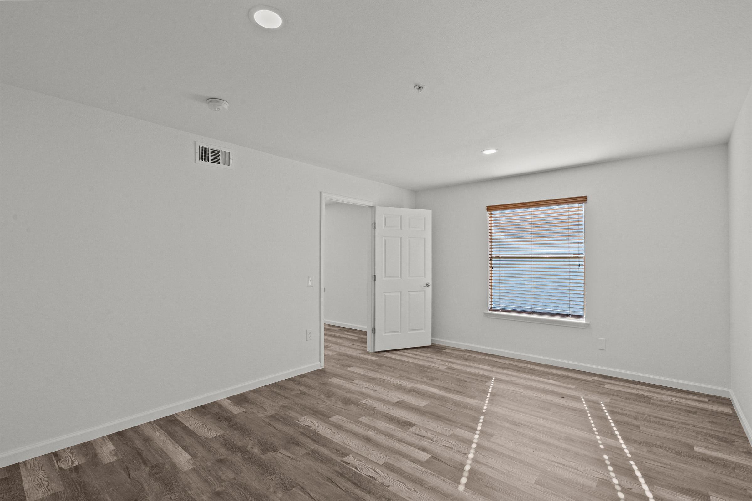 Interior view of a bright, empty room with light-colored walls, a wooden floor, and a large window with blinds. A door leads to an adjoining space, and recessed lighting provides illumination. The overall atmosphere is clean and spacious, ideal for various purposes.
