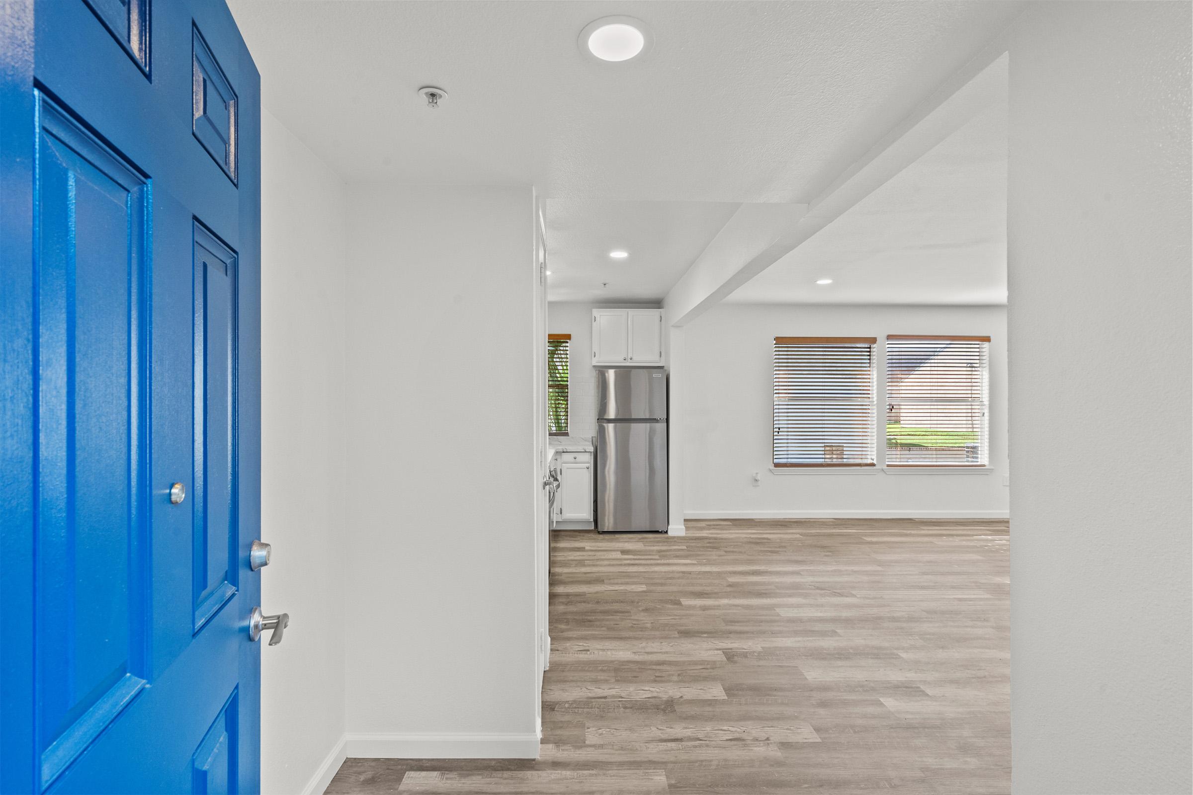 Bright entryway featuring a vibrant blue door, leading into an open concept living space with light-colored walls and wood flooring. The kitchen is visible in the background, equipped with stainless steel appliances. Large windows with blinds allow natural light to fill the area.