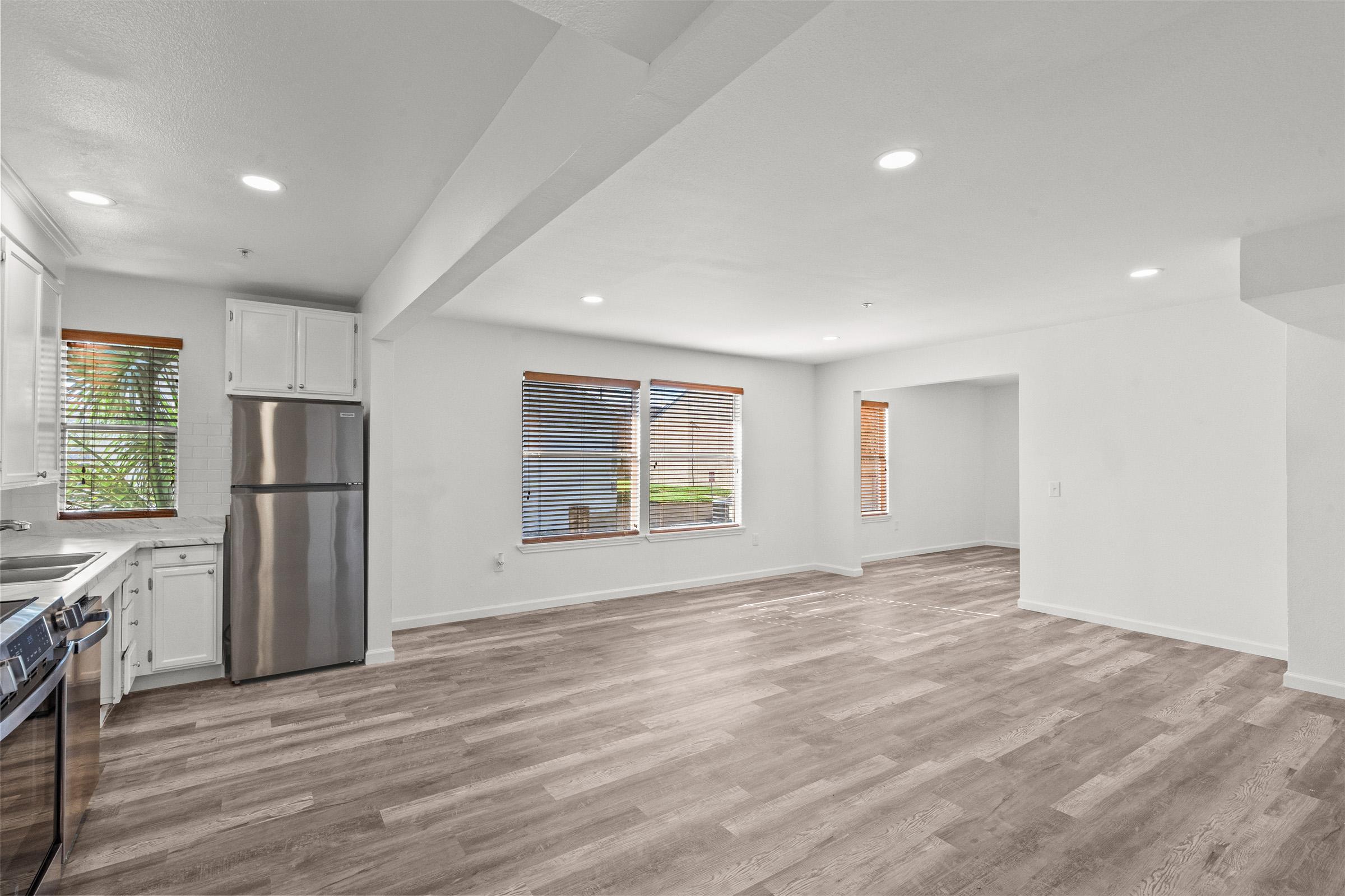A spacious, modern living area with light-colored walls and hardwood flooring. The room features large windows with horizontal blinds, allowing natural light to enter. A stainless steel refrigerator is visible in the corner, and the layout flows into an adjacent room, creating an open atmosphere.