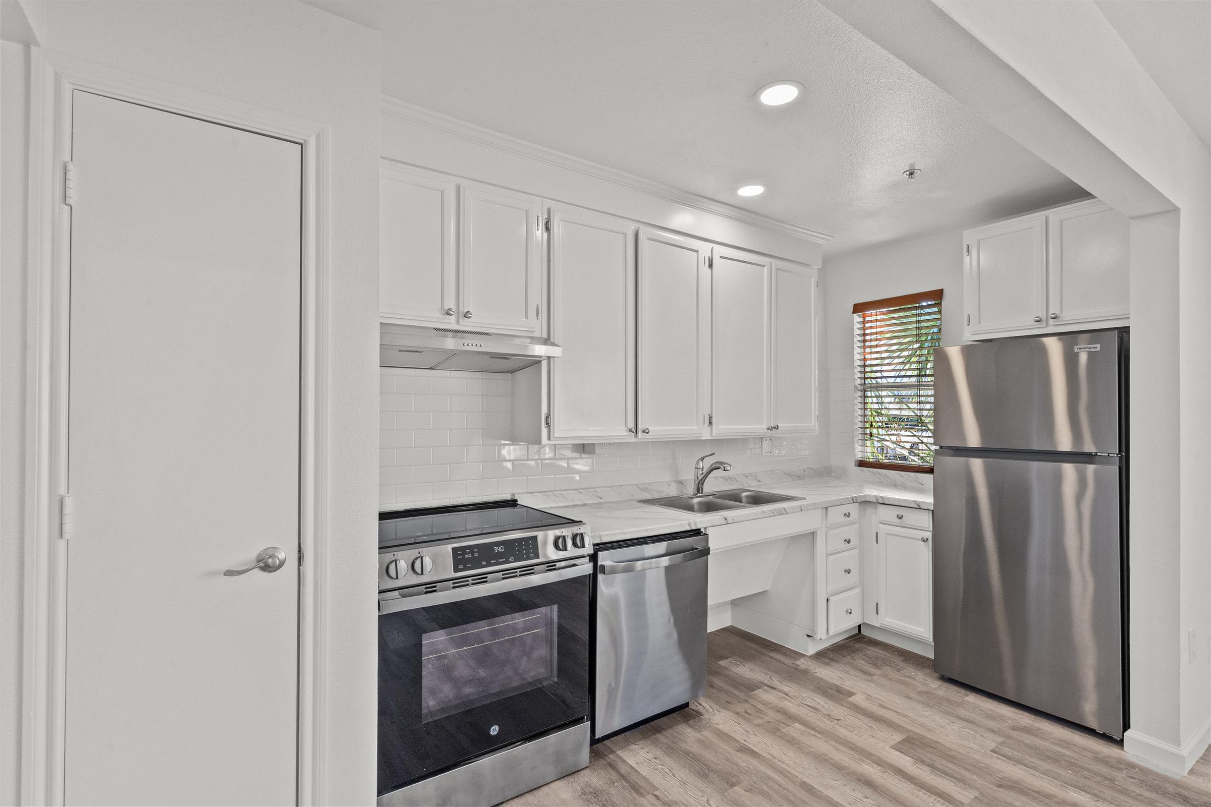 A modern kitchen featuring white cabinetry, stainless steel appliances including an oven, stove, and refrigerator, a sink, and a window with blinds. The flooring is light-colored wood, and the space is well-lit with overhead lighting.