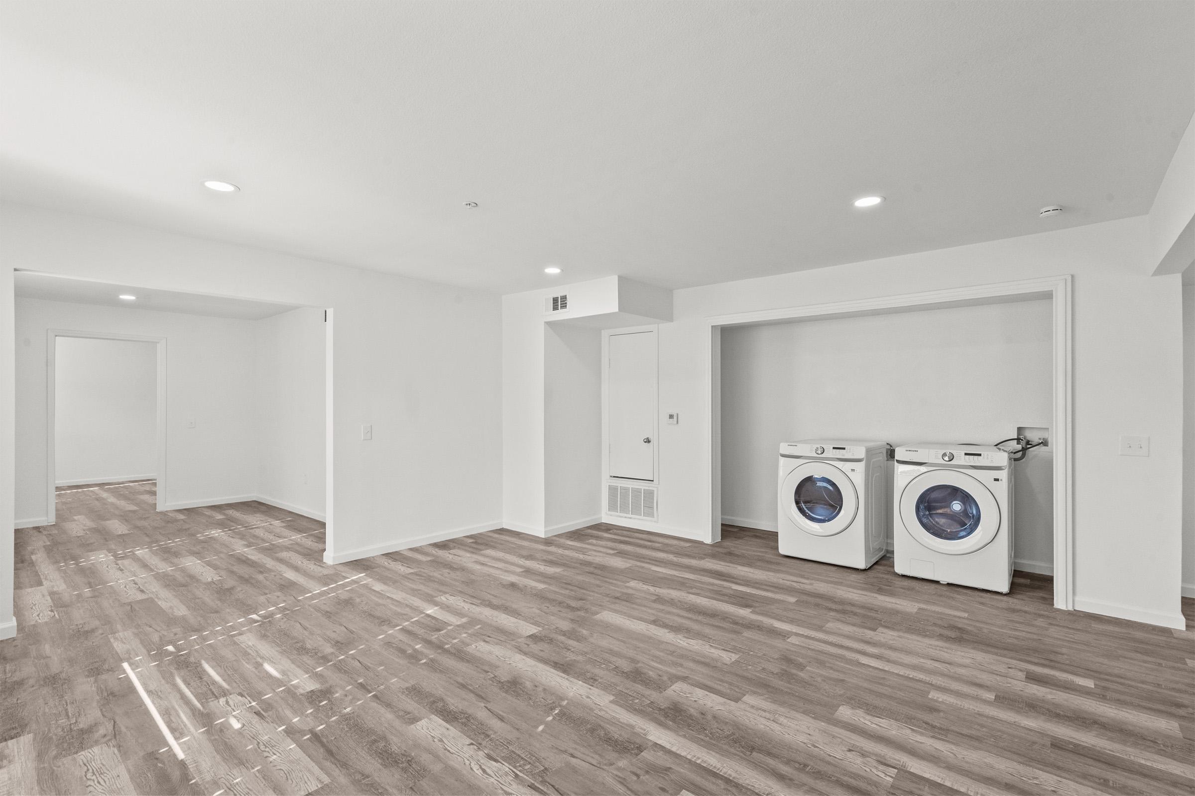 A spacious, empty room with light-colored walls and a wooden floor. Two white front-loading washing machines are visible in a built-in closet. The area is well-lit with recessed lighting, and there is a doorway leading to another room on the left.