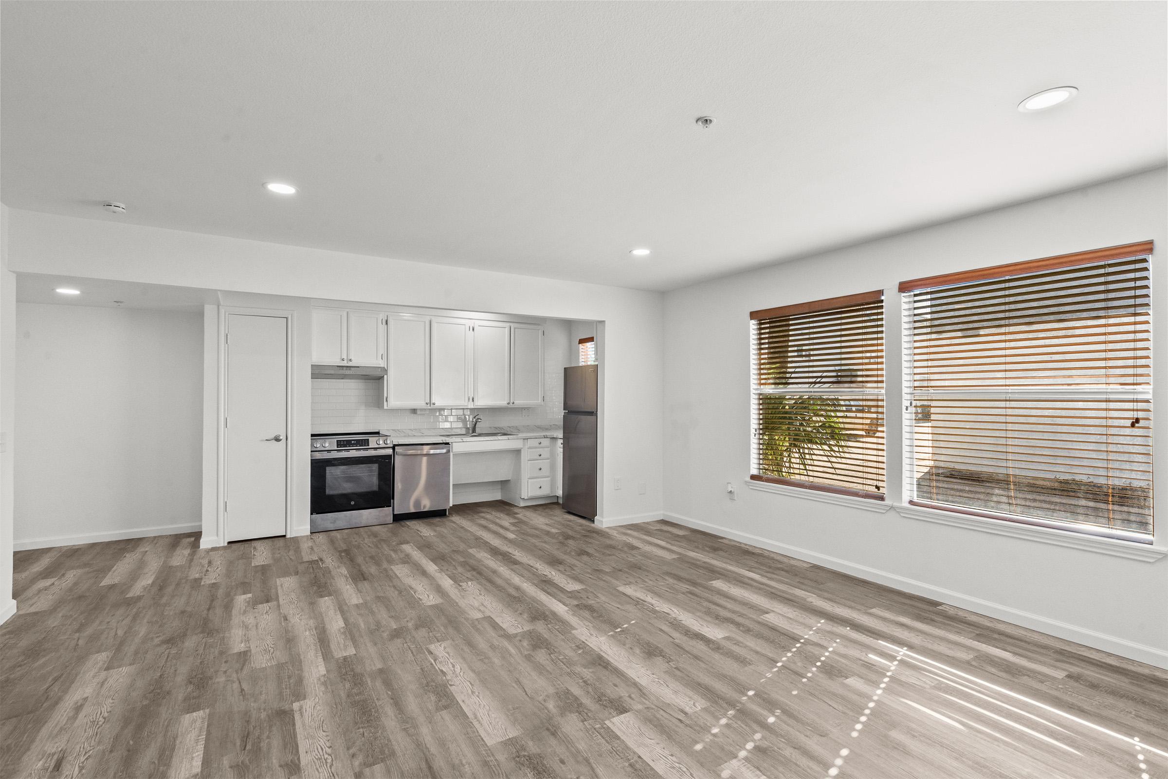 A modern, open-concept living space featuring light wood flooring, white walls, and ample natural light. The kitchen area includes stainless steel appliances and white cabinetry, while large windows with blinds provide views of the outdoors. The room appears spacious and inviting.