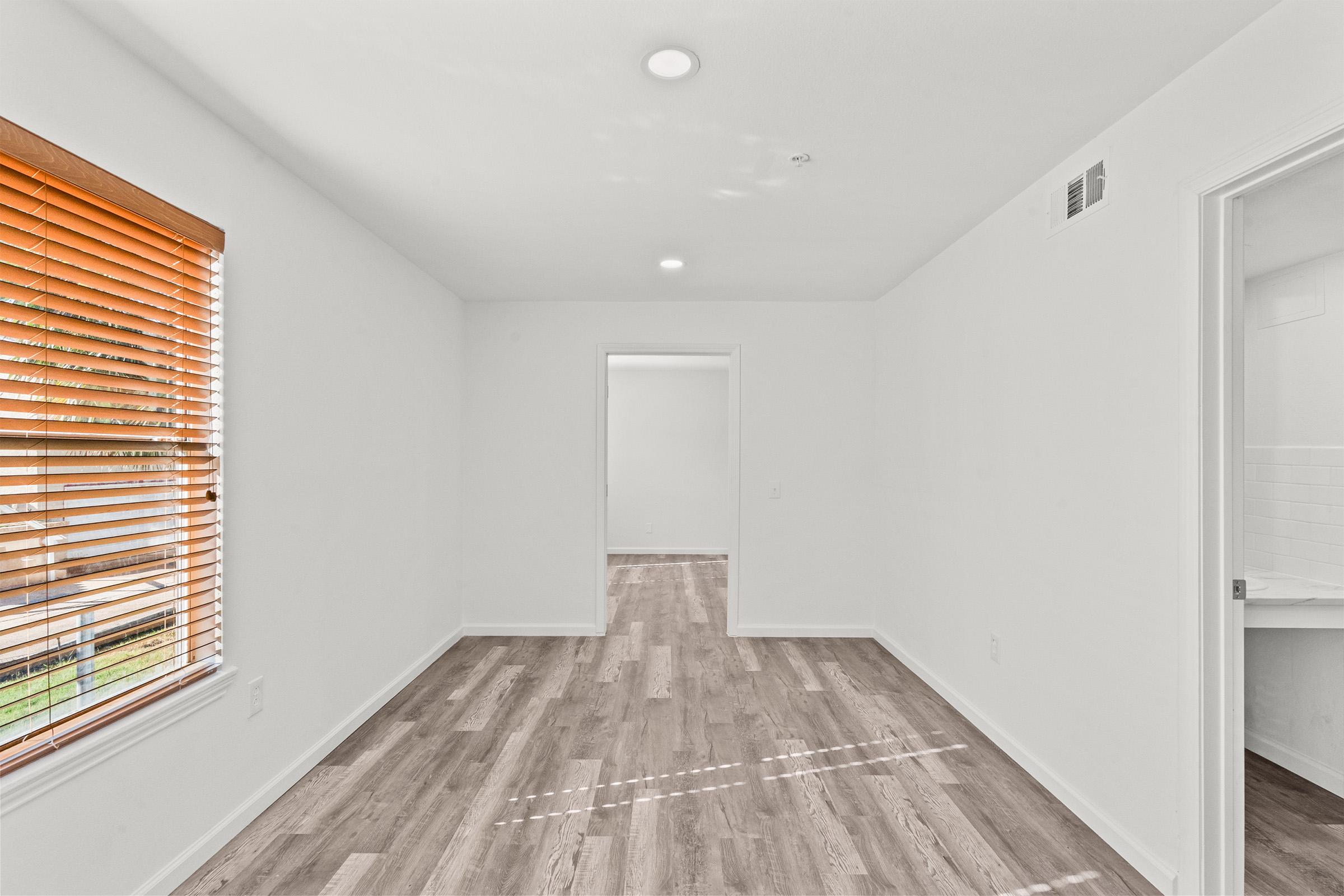 A bright, empty room with white walls and hardwood flooring. There are two light fixtures on the ceiling and a window with wooden blinds letting in natural light. A doorway leads to another room, and a bathroom is partially visible in the corner. The space appears clean and well-maintained.