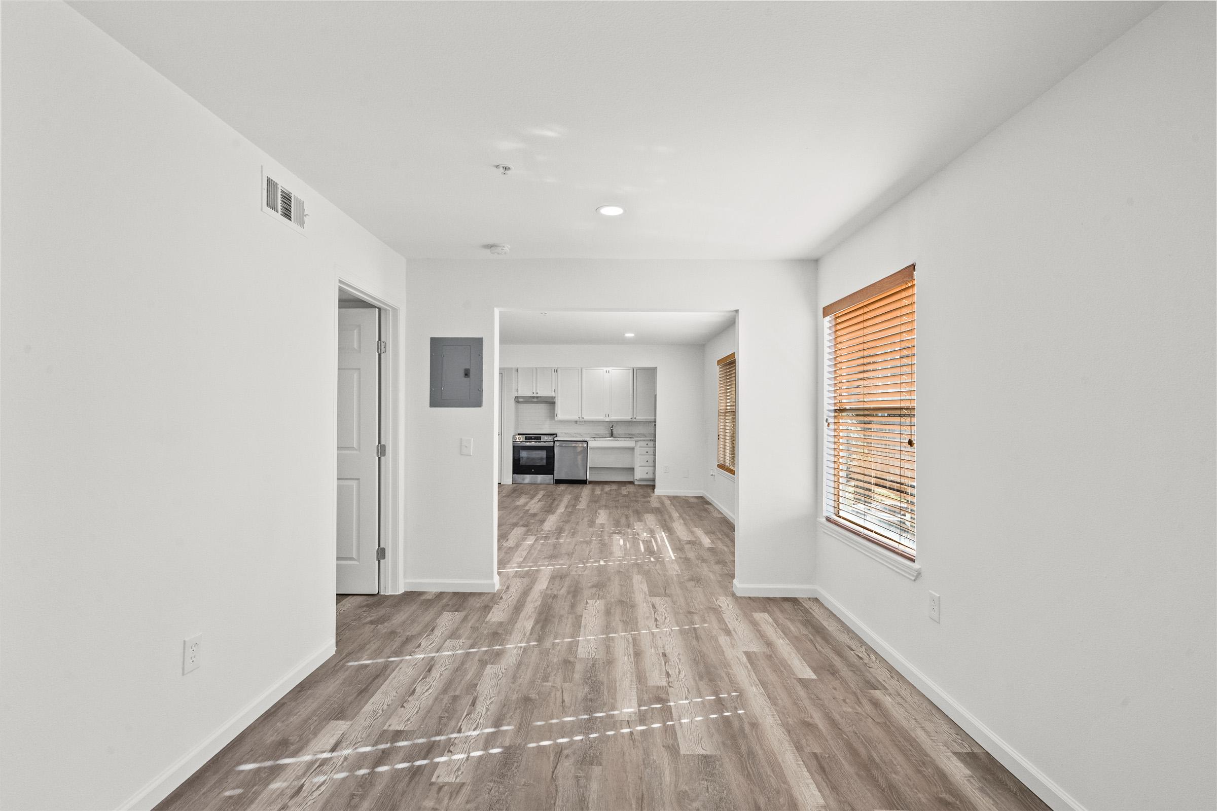 A bright, modern room featuring light-colored walls and a spacious layout. There are wood-like flooring and large windows with blinds. In the background, a kitchen area is visible with white cabinetry and stainless steel appliances. A door leads to another room, enhancing the open concept feel.
