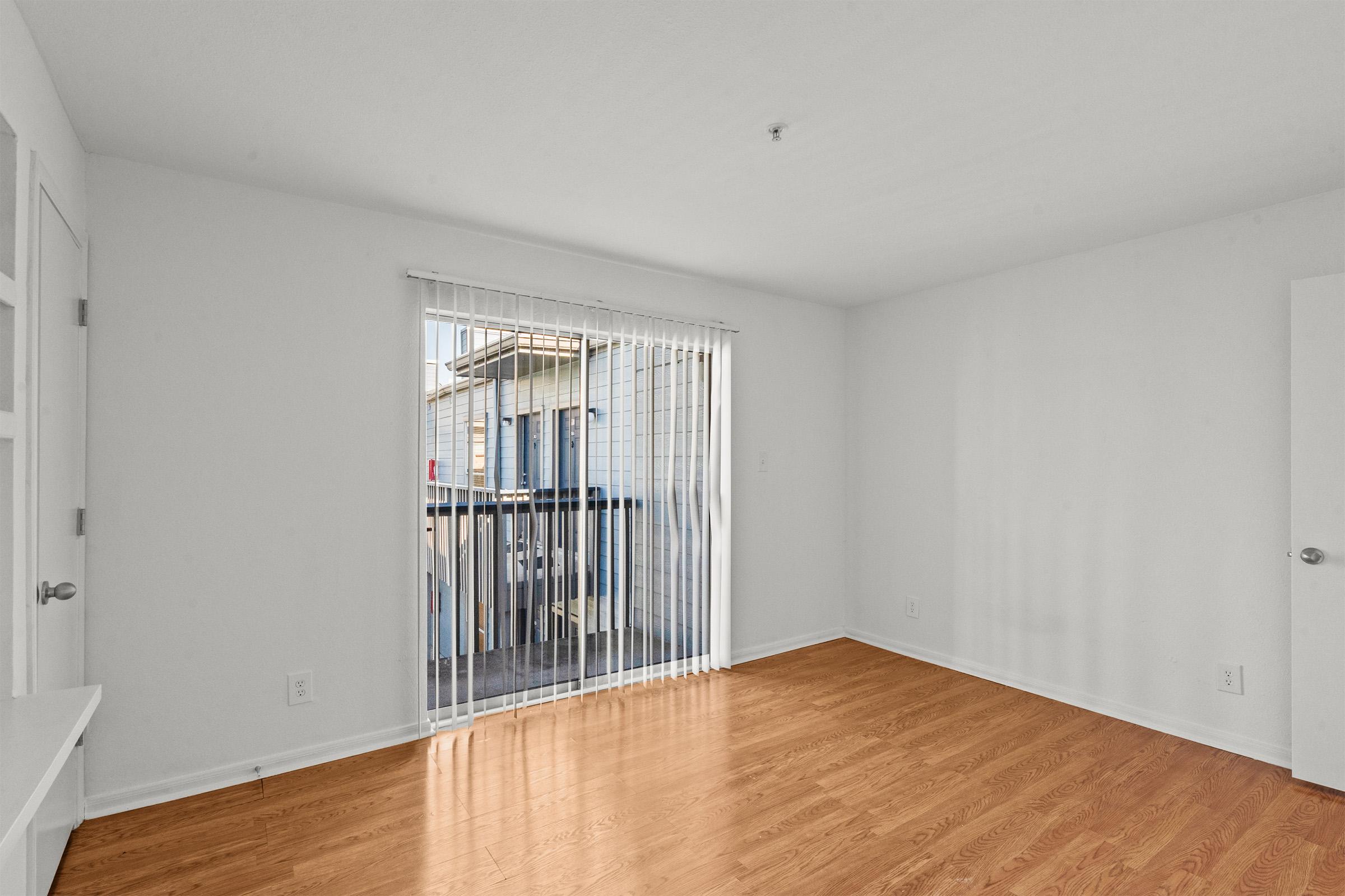 Empty room with light-colored walls and a wooden floor. A large window with vertical blinds allows natural light to enter, revealing an outdoor balcony area. The space is minimalistic, featuring a door and a small wall area that could accommodate furniture.
