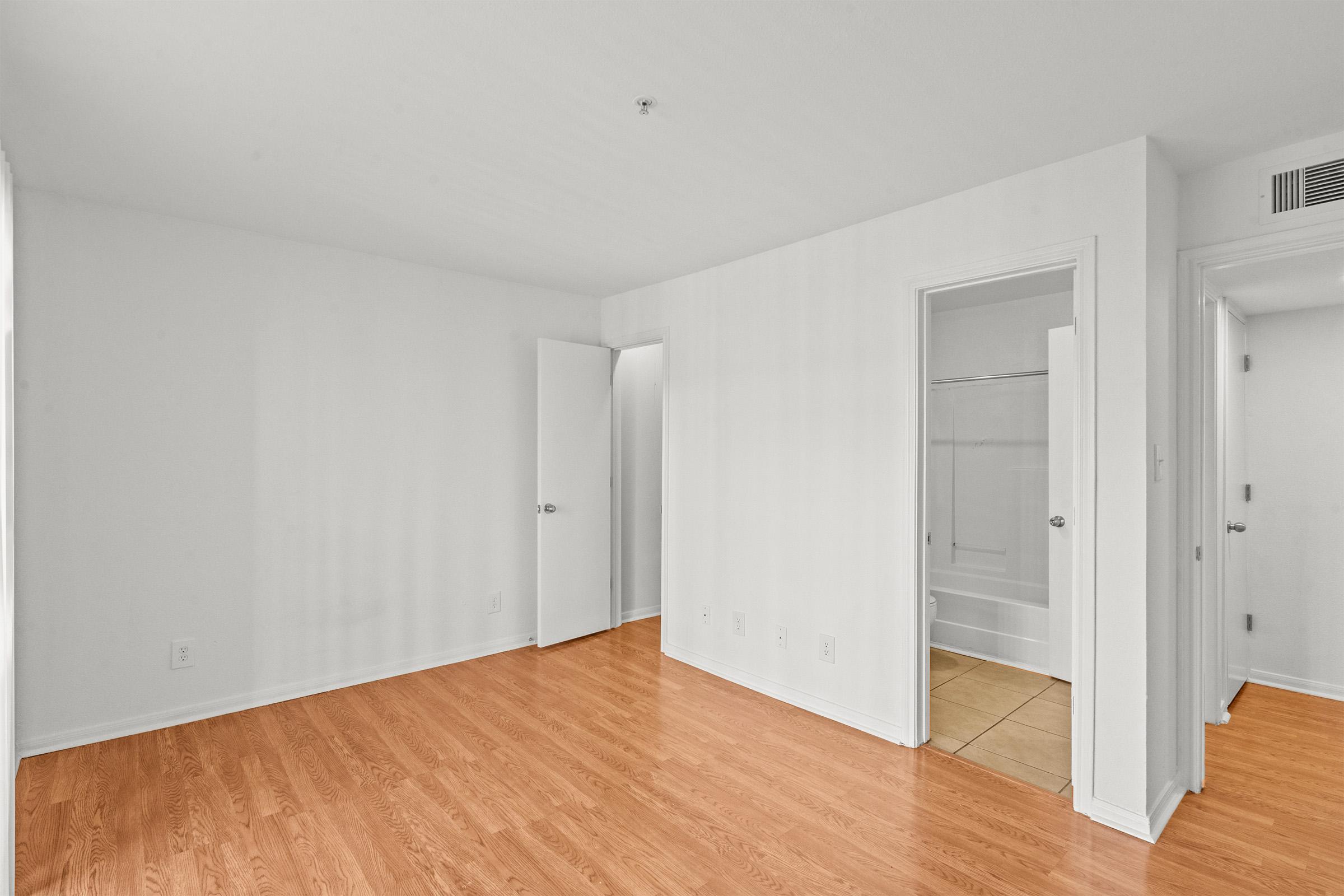Empty room featuring hardwood flooring, white walls, and doors leading to adjacent spaces. Natural light enters through a window. The room has a simple layout with no furniture, showcasing a clean and minimalist aesthetic. The bathroom doorway is visible in the background.
