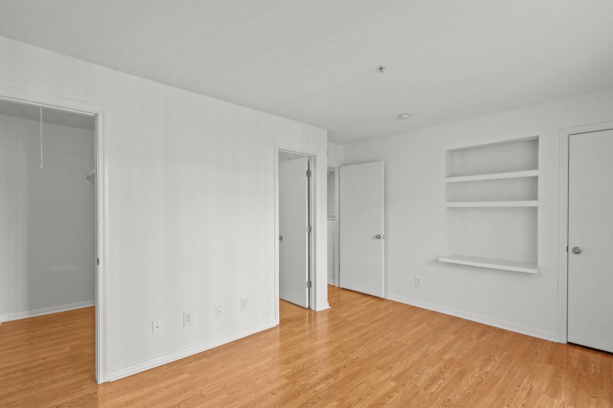 A spacious, empty room with light-colored walls and a wooden floor. There are two closed doors, one leading to another room and the other to a closet. A small wall shelf is mounted on the right side, adding a subtle decorative element to the otherwise minimalist space. Natural light fills the area, creating an airy feel.