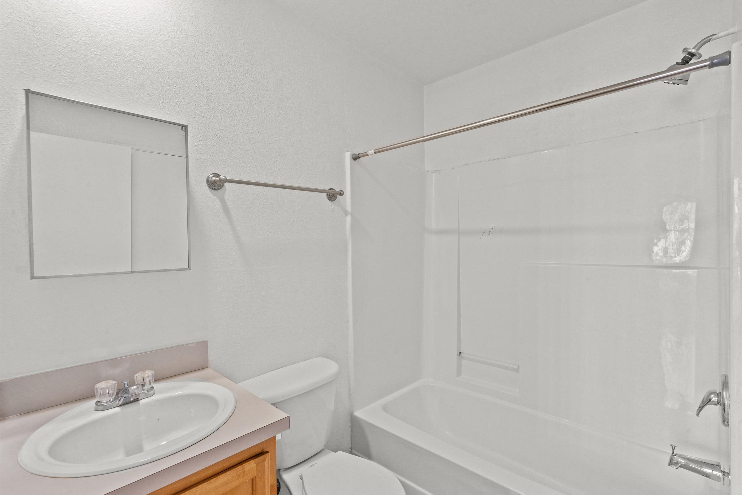 A clean, modern bathroom featuring a white bathtub, a toilet, and a sink with a simple countertop. The walls are painted white, and there is a large mirror above the sink. A shower rod is installed above the bathtub, completing the uncluttered and bright appearance of the space.
