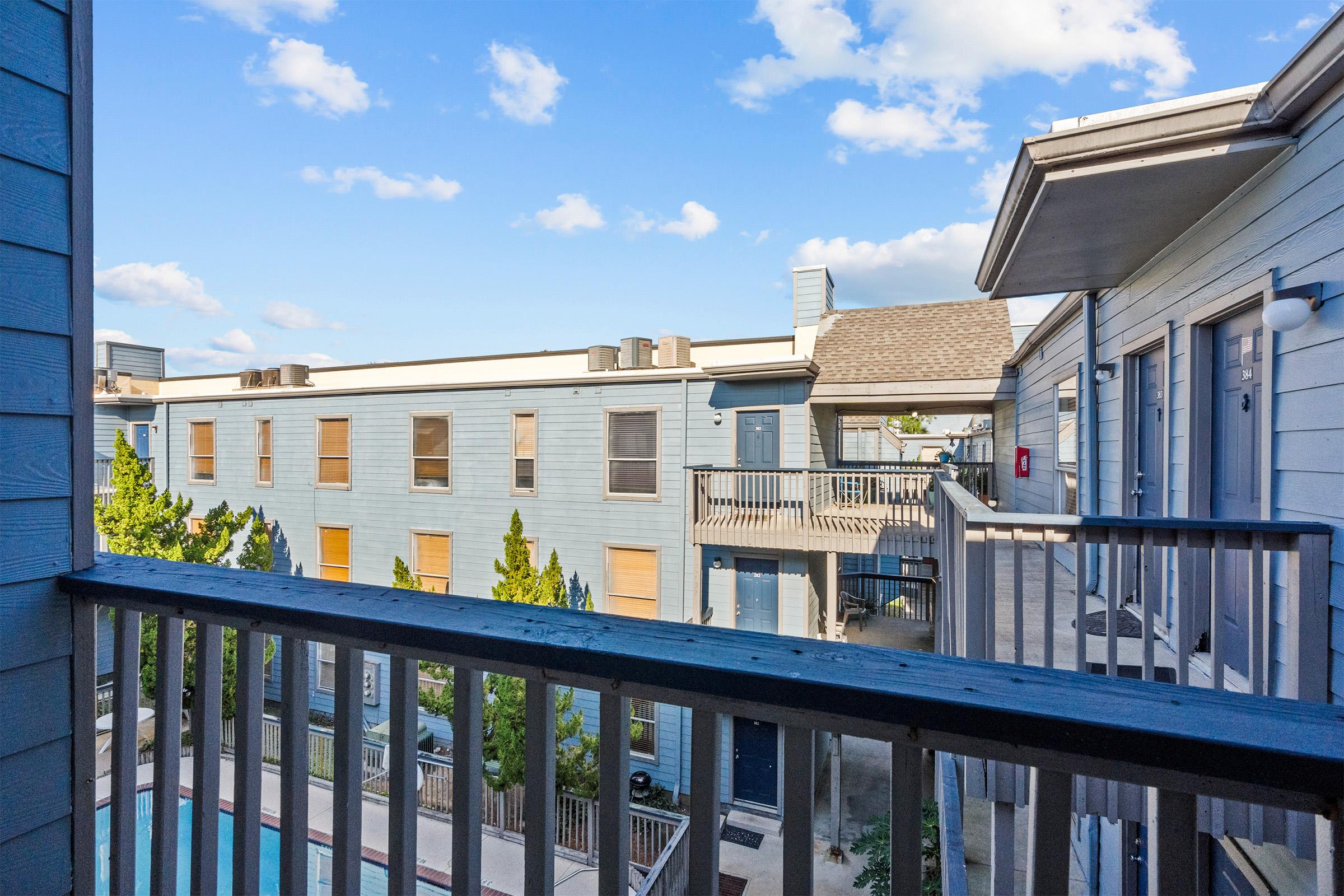 View from a balcony overlooking a courtyard with a pool and buildings. The scene includes blue sky and clouds, with several trees in the foreground and multiple apartment units visible, featuring balconies and windows. The setting conveys a tranquil residential atmosphere.