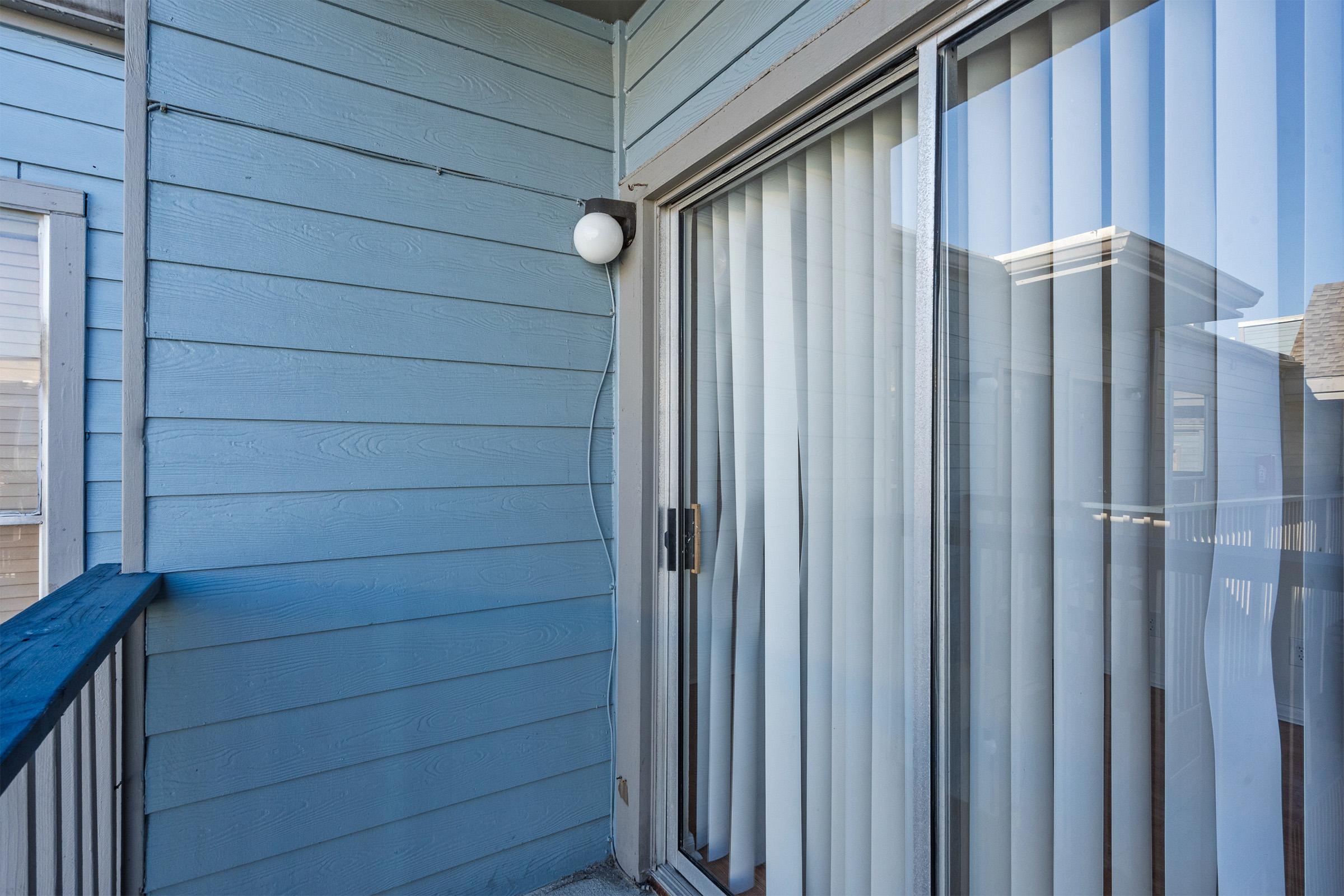 A balcony with light fixture and sliding glass doors, featuring vertical blinds. The walls are painted light blue, and there are wooden panels along the edge of the balcony. Two windows are visible, contributing to a bright and airy atmosphere.