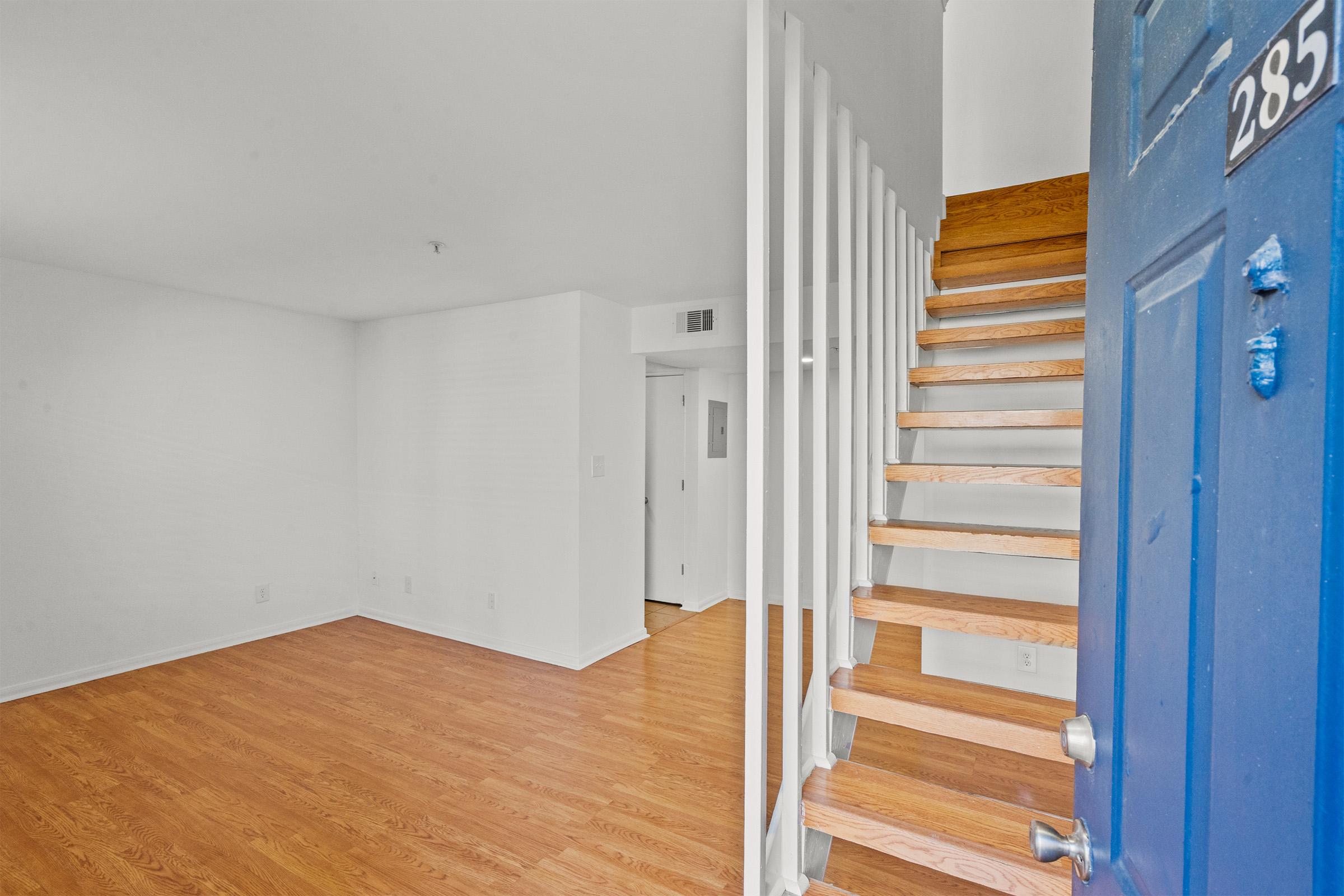 Interior view of a bright, empty room with light wooden flooring. A staircase with wooden steps leads to an upper level on the right. The walls are painted white, and there's a blue front door visible on the left side of the image. Ideal for a rental or new home space.