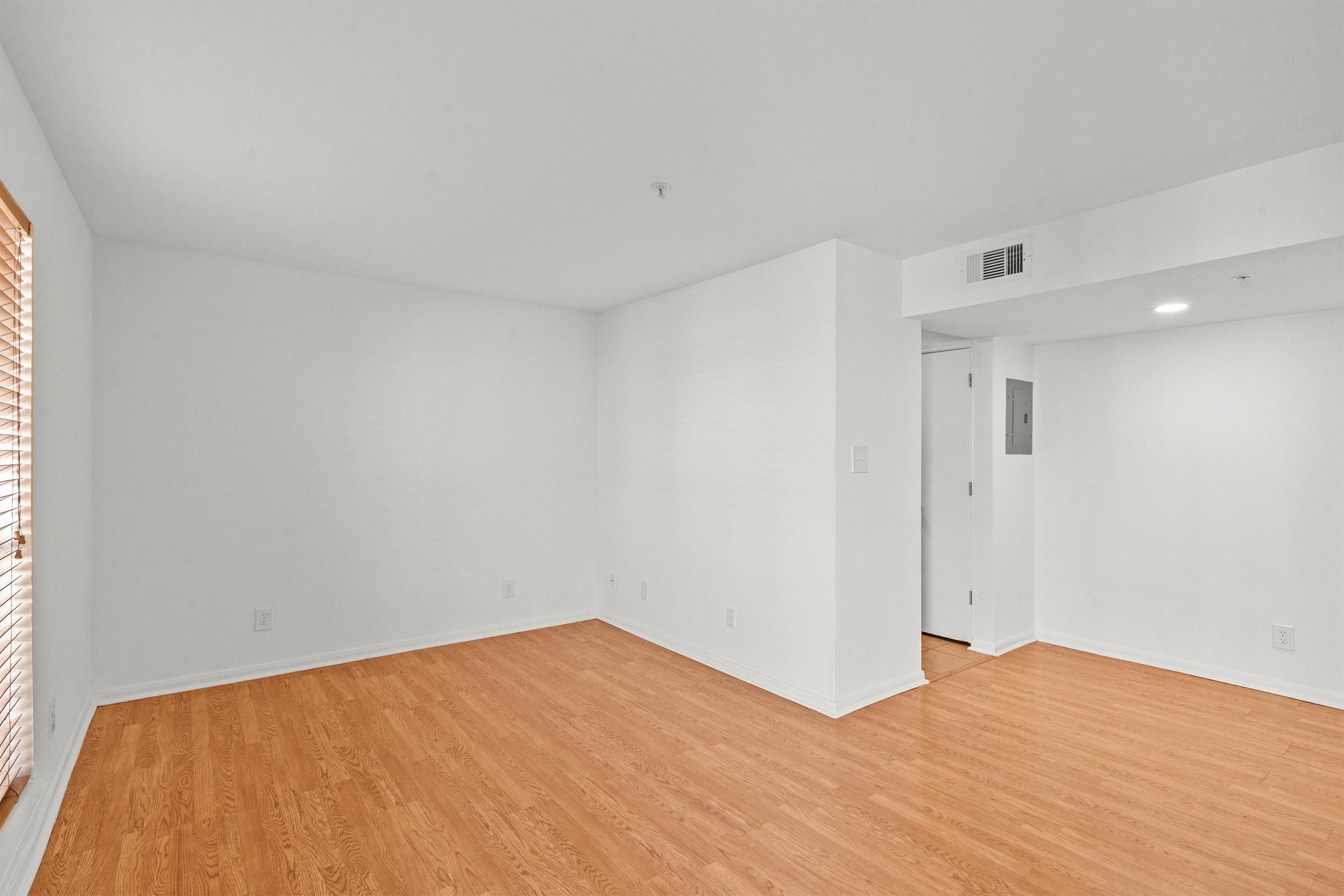 A bright, empty room with light-colored hardwood floors and white walls. A window with blinds allows natural light to enter, and a door is visible on the right side leading to another space. The room appears spacious and minimalistic, suitable for various design options.