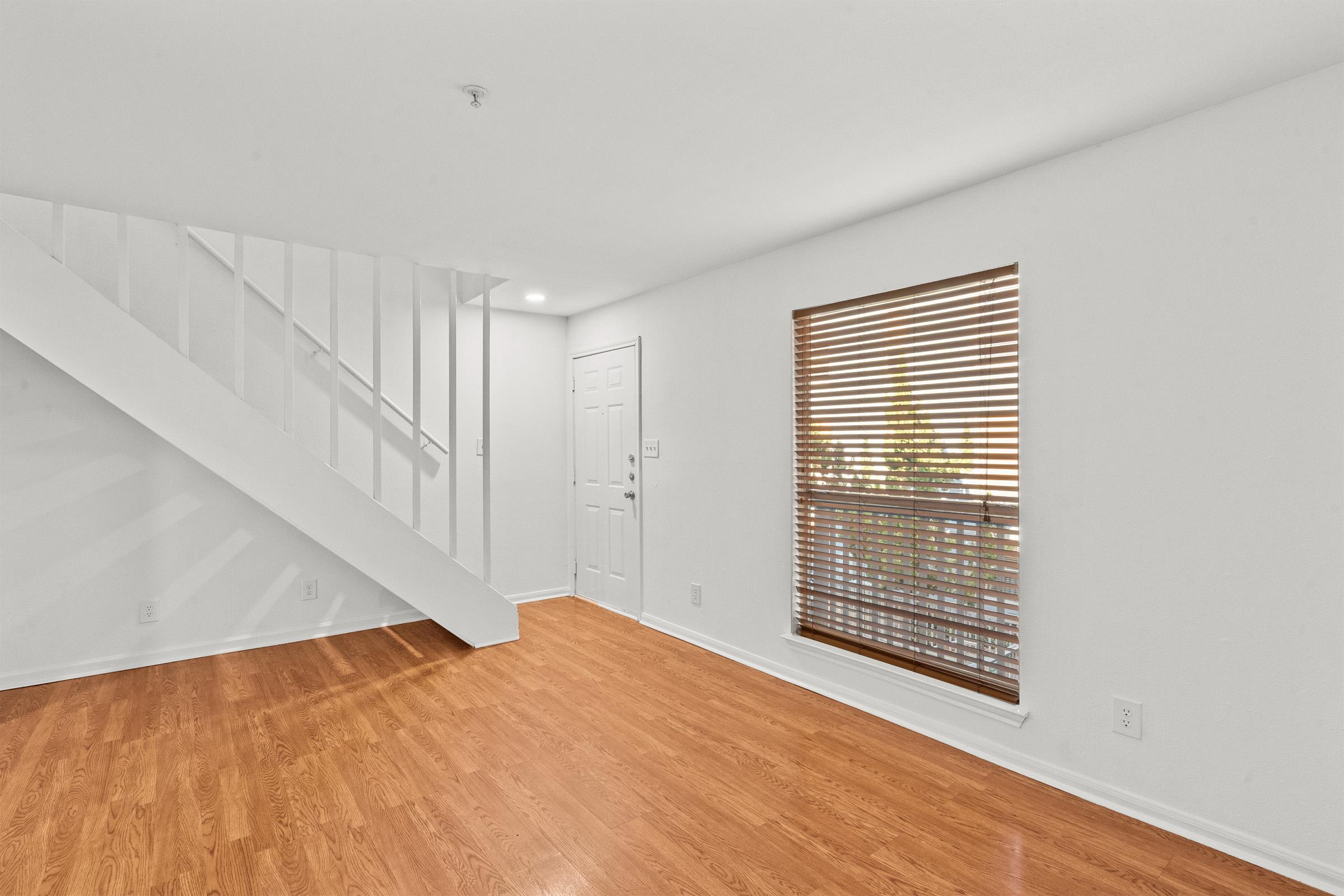 Interior view of a bright, empty room with light-colored walls and hardwood flooring. A staircase is visible on the left, and a large window with wooden blinds lets in natural light. The main door is located at the far end, creating a welcoming and spacious atmosphere.