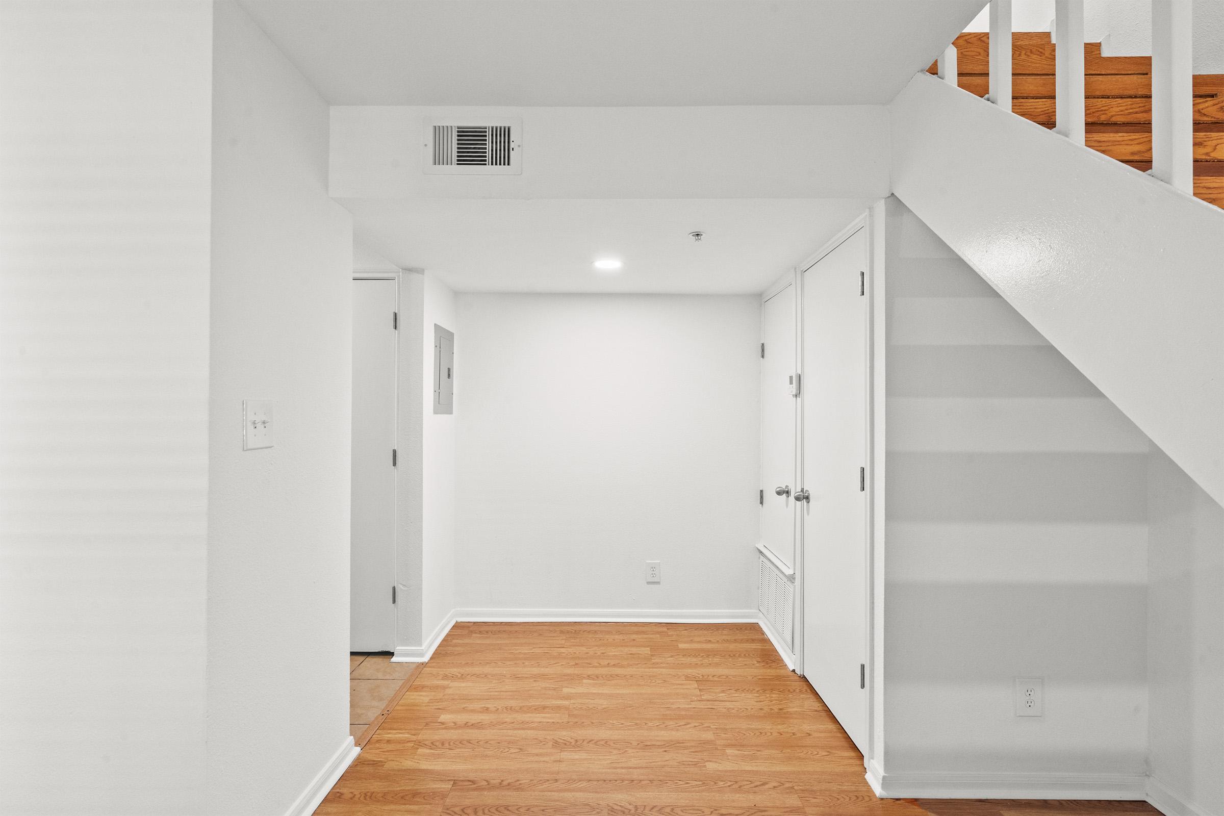 Interior view of a bright hallway featuring white walls and wooden flooring. On the left, there's a door leading to a room, while another door can be seen further down the hall. A staircase leading to an upper level is visible on the right side, adding depth to the space. The area is well-lit and clean.