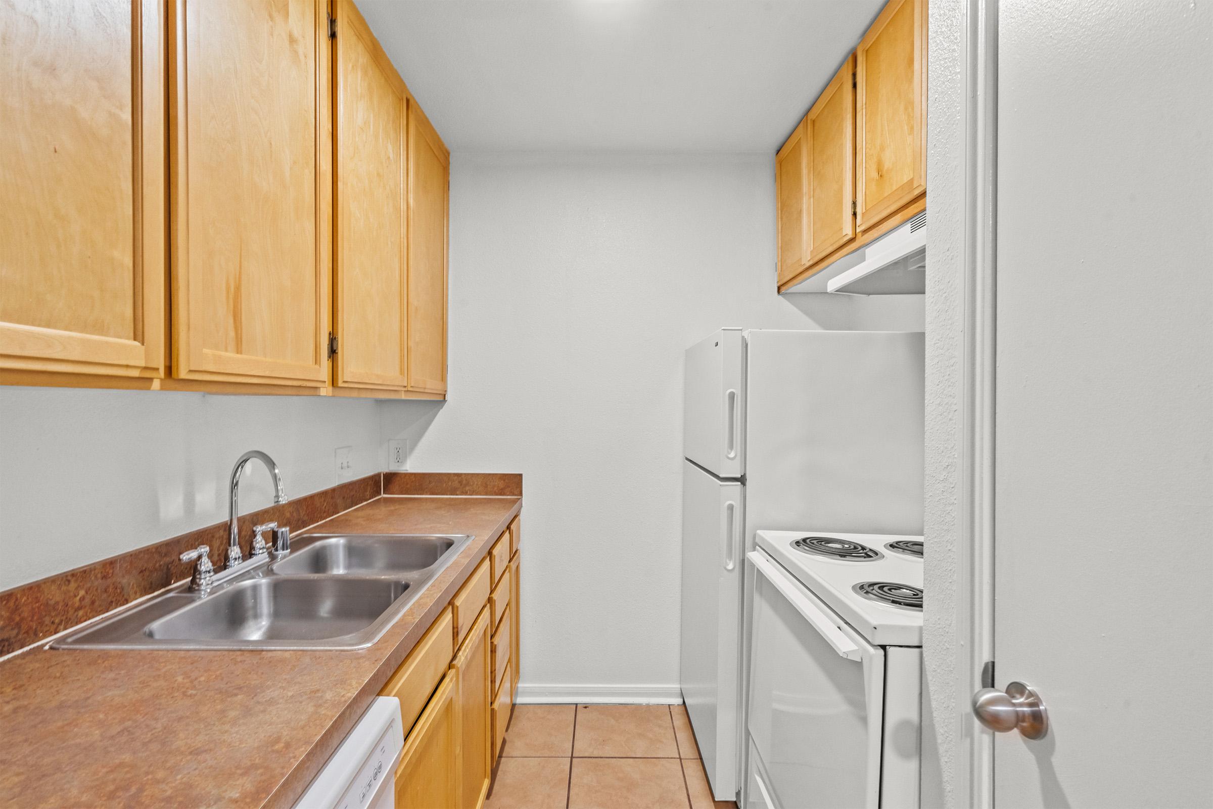 A small kitchen featuring light wood cabinets, a countertop with a double sink, a white refrigerator, and a white stove with an oven. The walls are painted in a neutral color, and the floor is tiled. The layout is compact and functional for basic cooking needs.