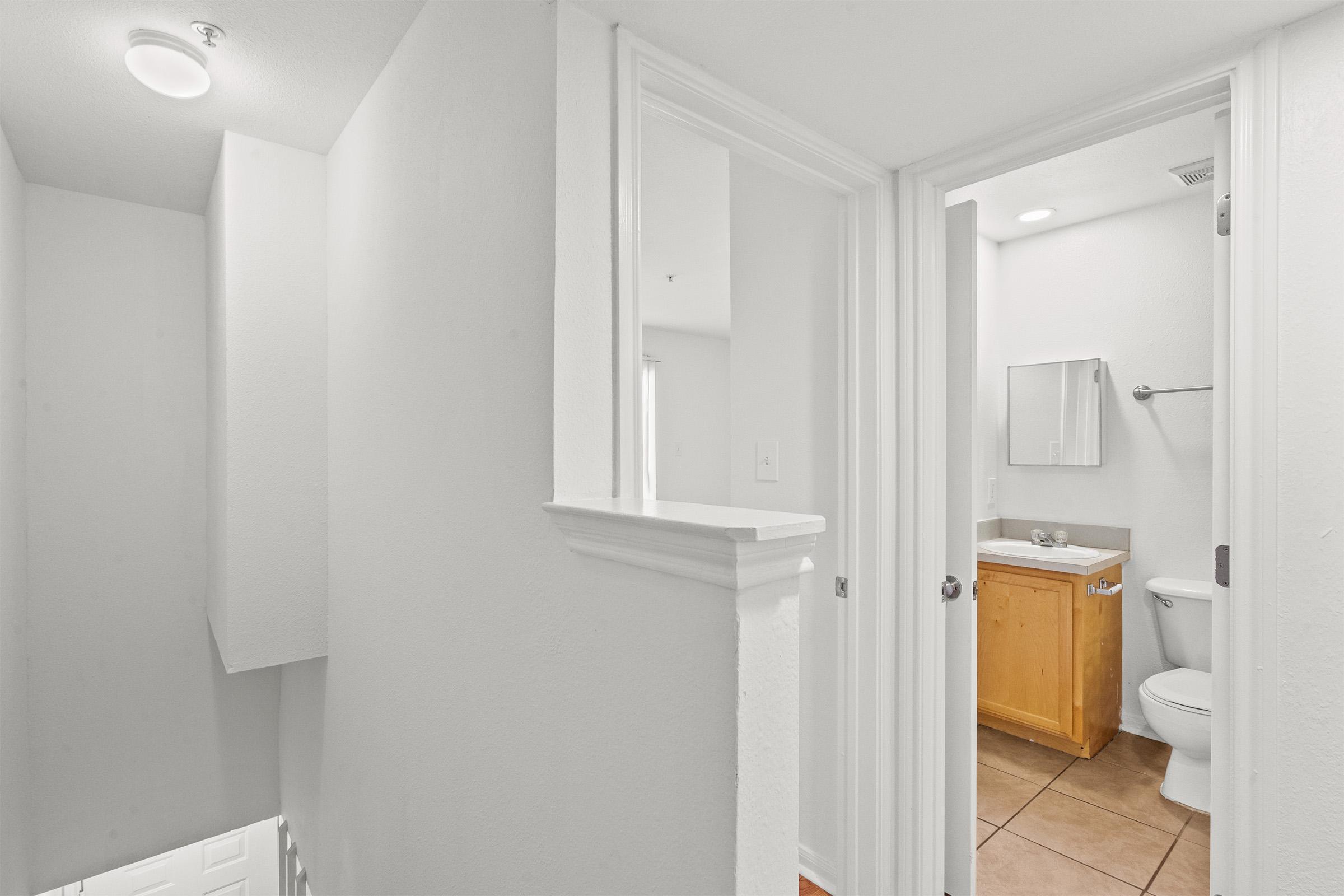 A bright, clean hallway leading to a bathroom. The hallway features white walls and a ceiling light. On the right, a bathroom is visible with a wooden cabinet, sink, mirror, and toilet. The flooring is tiled. Stairs are seen at the end of the hallway, leading to another level.