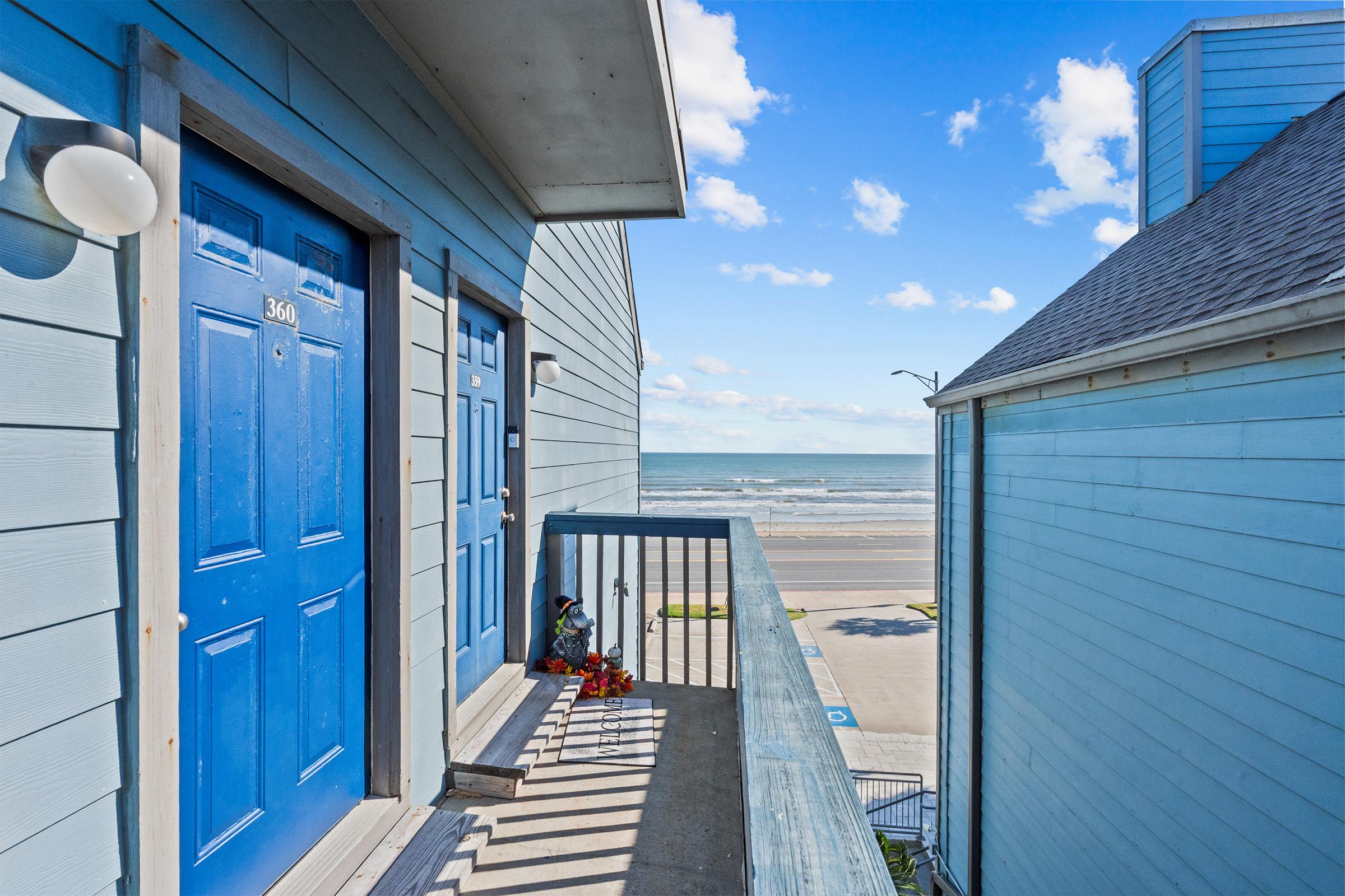 A view from a balcony overlooking the ocean. The scene includes two blue exterior doors with the left door numbered 3640. The boardwalk leads to the beach, visible in the distance under a clear sky with scattered clouds. A small decorative item and flowers are placed on the balcony railing.