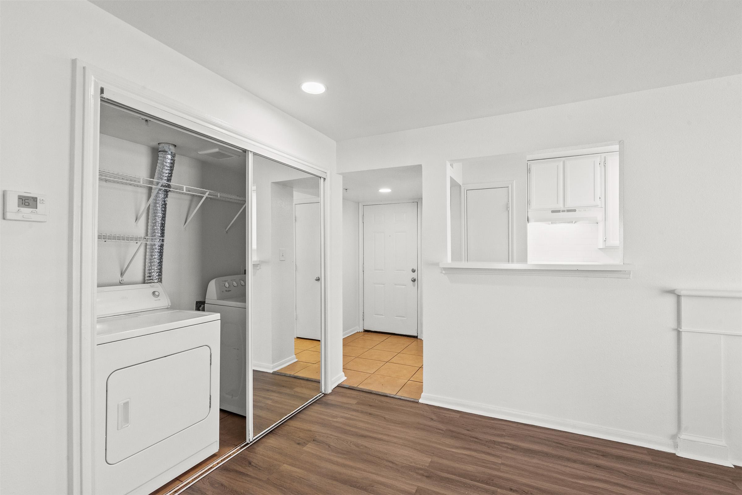 A bright, modern laundry area featuring a stacked washer and dryer, with a mirrored closet beside it. The room has light-colored walls and a seamless transition to a hallway with a tile floor. A small opening leads to another room, enhancing the open feel of the space.