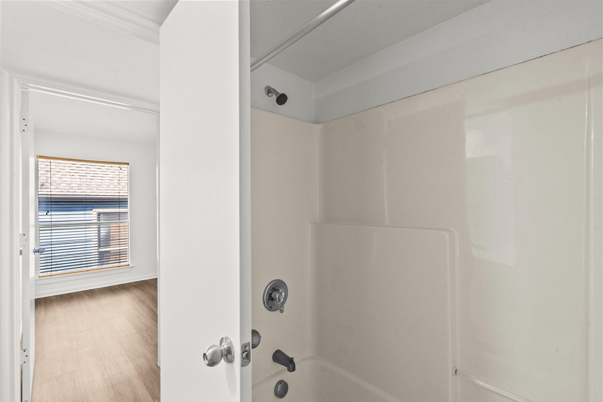 A view of a bathroom featuring a bathtub with a showerhead installed, with a light-colored wall and smooth surface. In the background, a window can be seen with blinds, allowing natural light to enter the space. The flooring appears to be a light wood finish. The overall ambiance is clean and modern.
