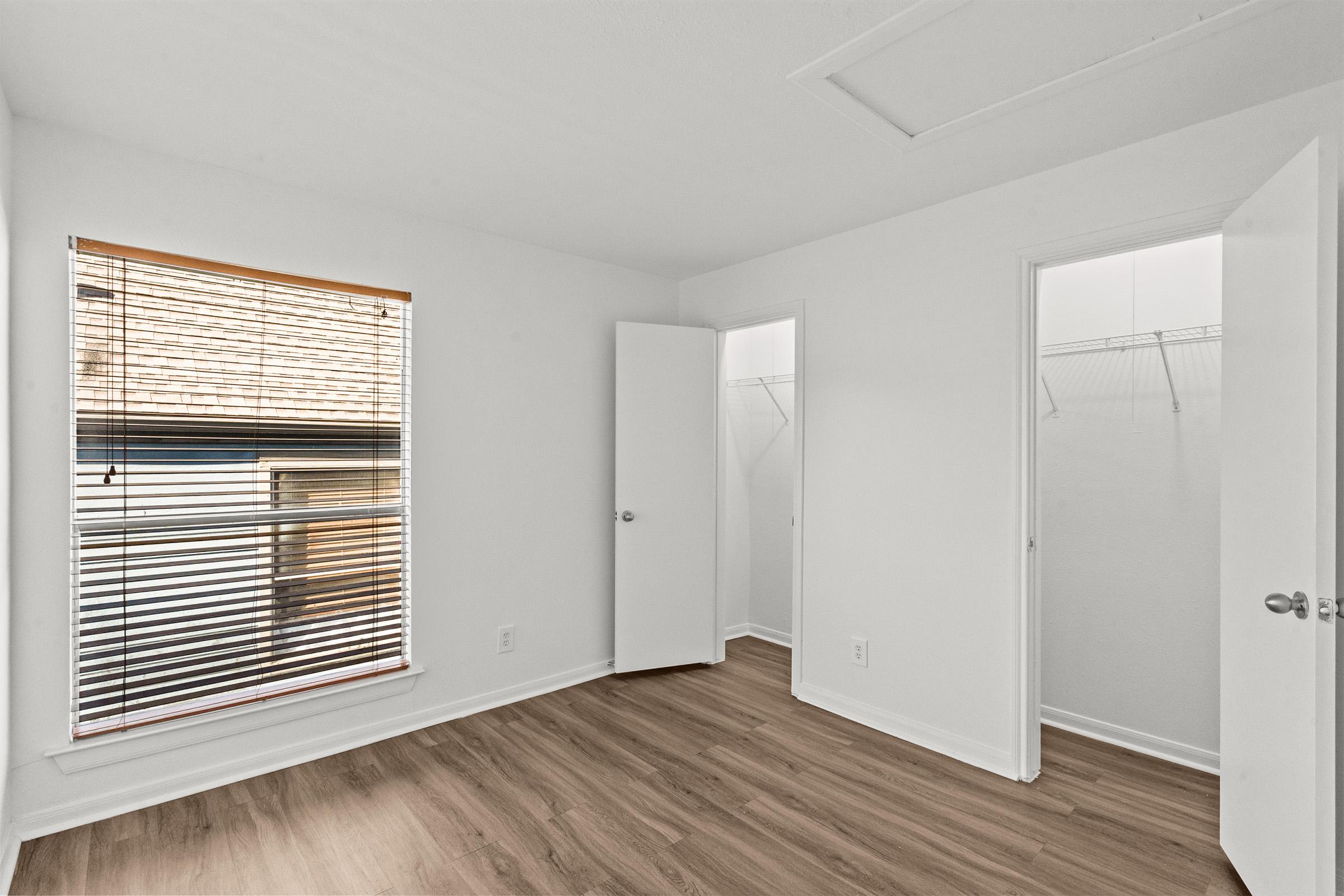 A bright, empty room with light-colored walls and wooden flooring. There is a window with blinds letting in natural light, a closet with an open door on the right, and another door leading to a small space. The overall atmosphere is clean and spacious, ideal for personal decoration.