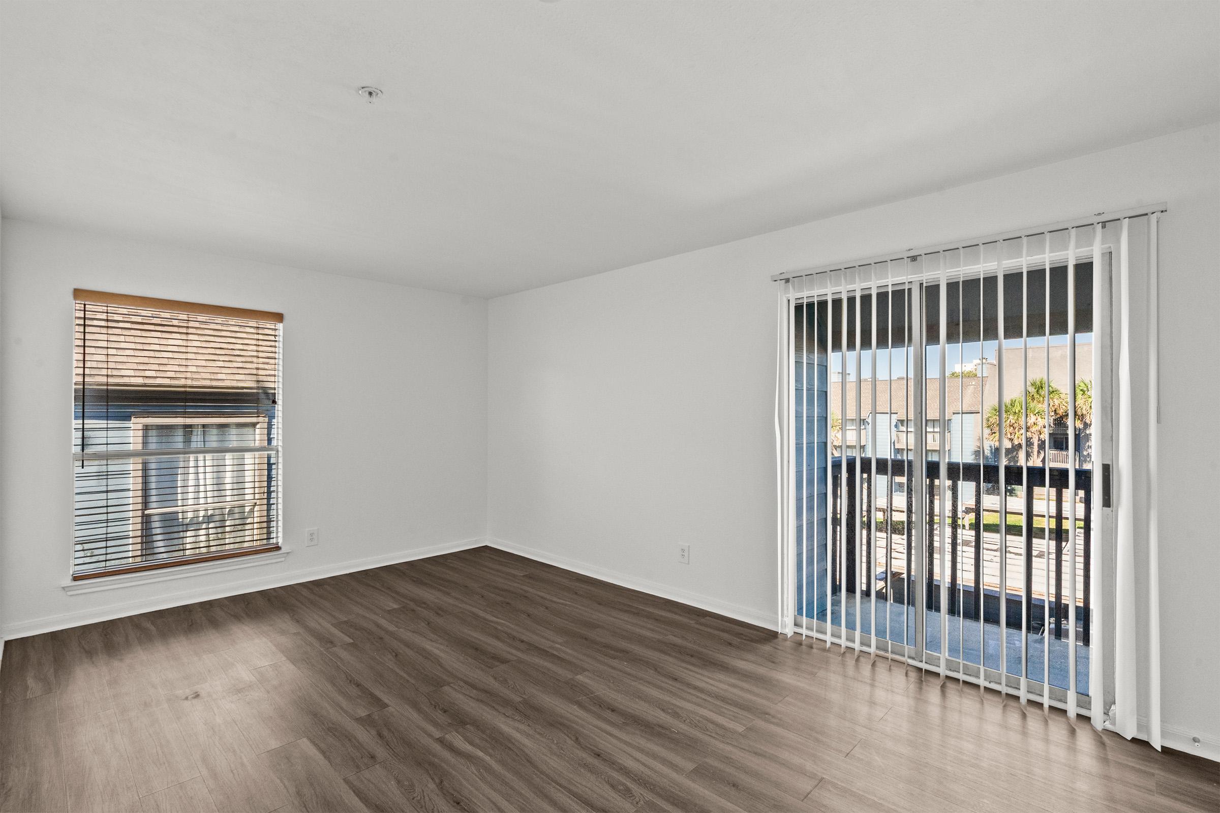 Bright, empty room with light-colored walls and hardwood flooring. A window with blinds lets in natural light, and there's a glass door with vertical blinds leading to a balcony. The space is clean and unfurnished, suggesting potential for interior design.