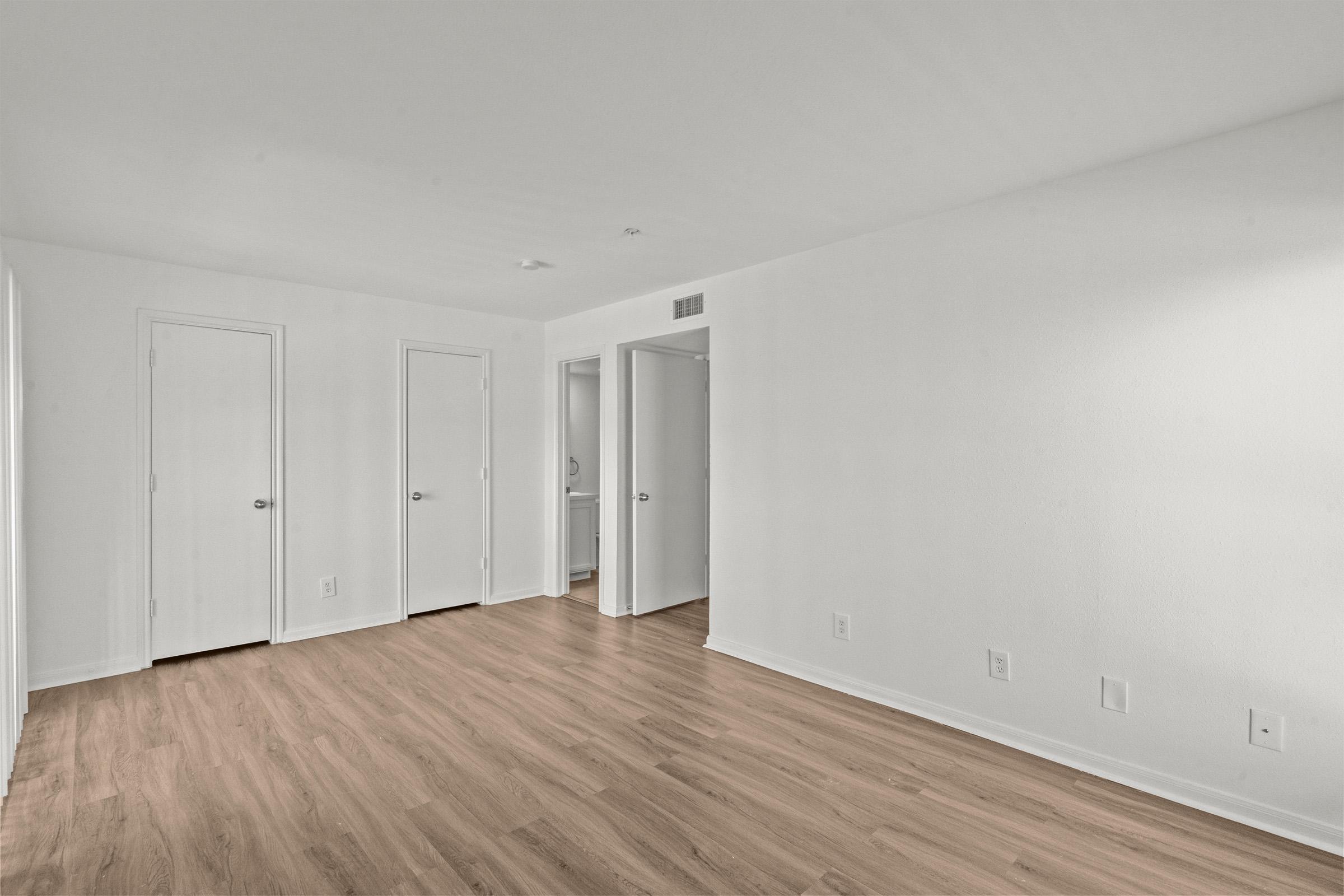 A bright, empty room with light-colored walls and wooden flooring. There are two closed doors on the left and one open door on the right leading to another room. The space is well-lit, creating a spacious and clean ambiance.