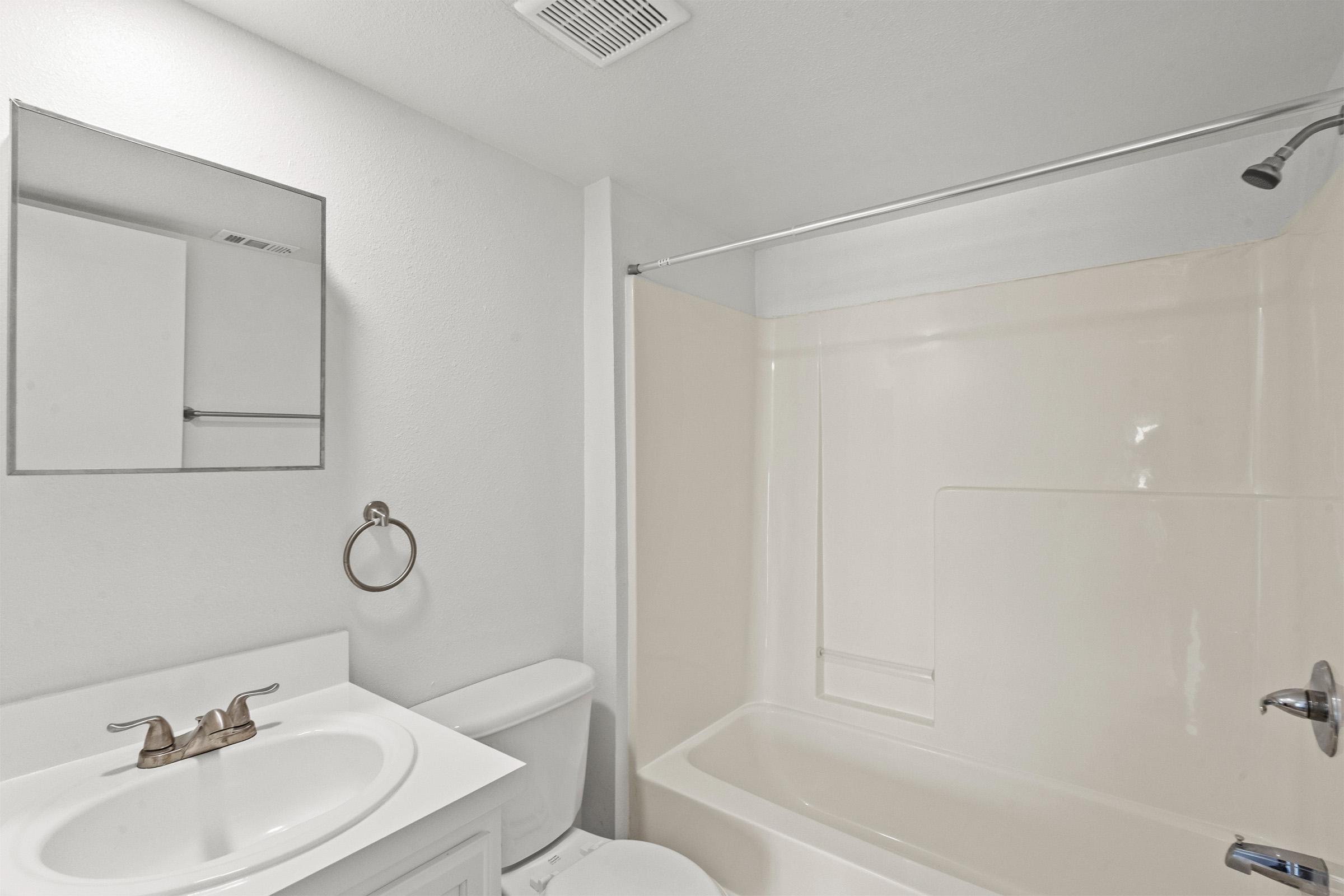 A clean, modern bathroom featuring a white bathtub with a shower curtain, a sink with brushed nickel faucets, a round mirror above the sink, and a toilet. The walls and fixtures are primarily white, creating a bright and spacious feel.