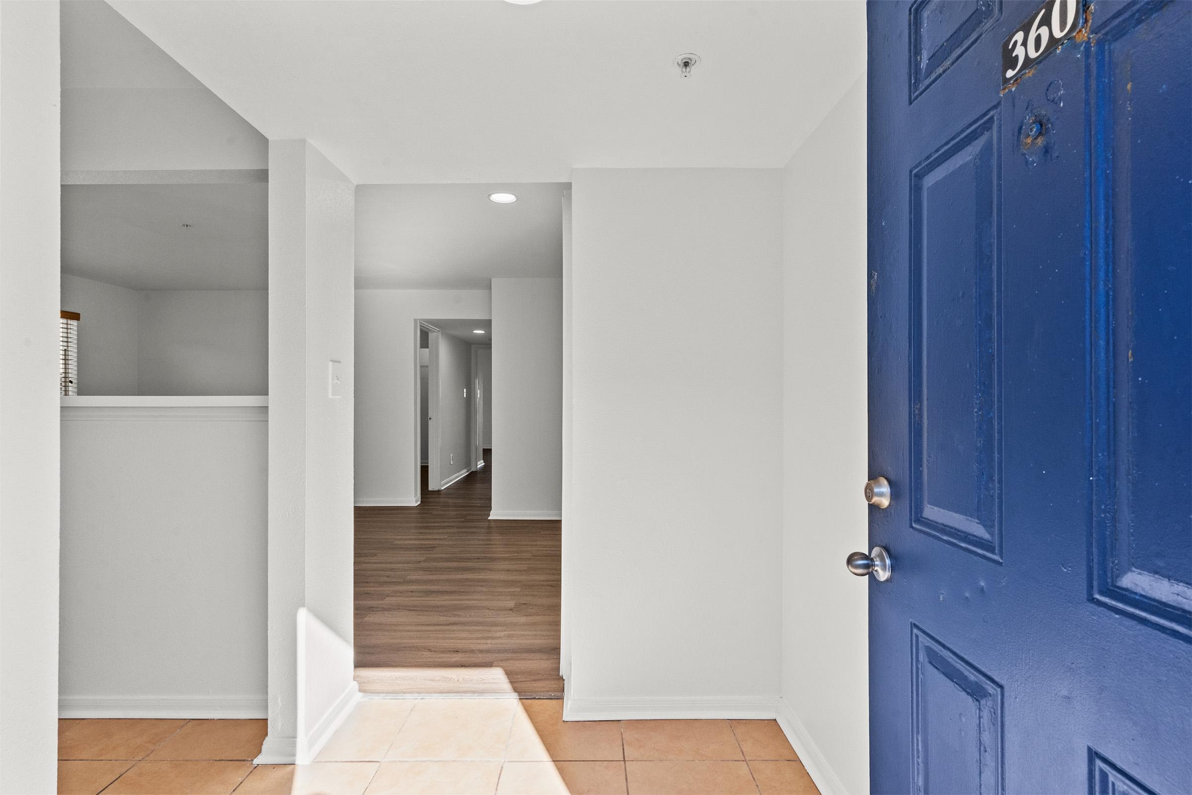 Interior view of an entrance hall featuring a blue front door. The flooring includes tiled and wooden sections. A hallway leads deeper into the space, with bright lighting and white walls. There is an opening to the left, possibly leading to another room or area.