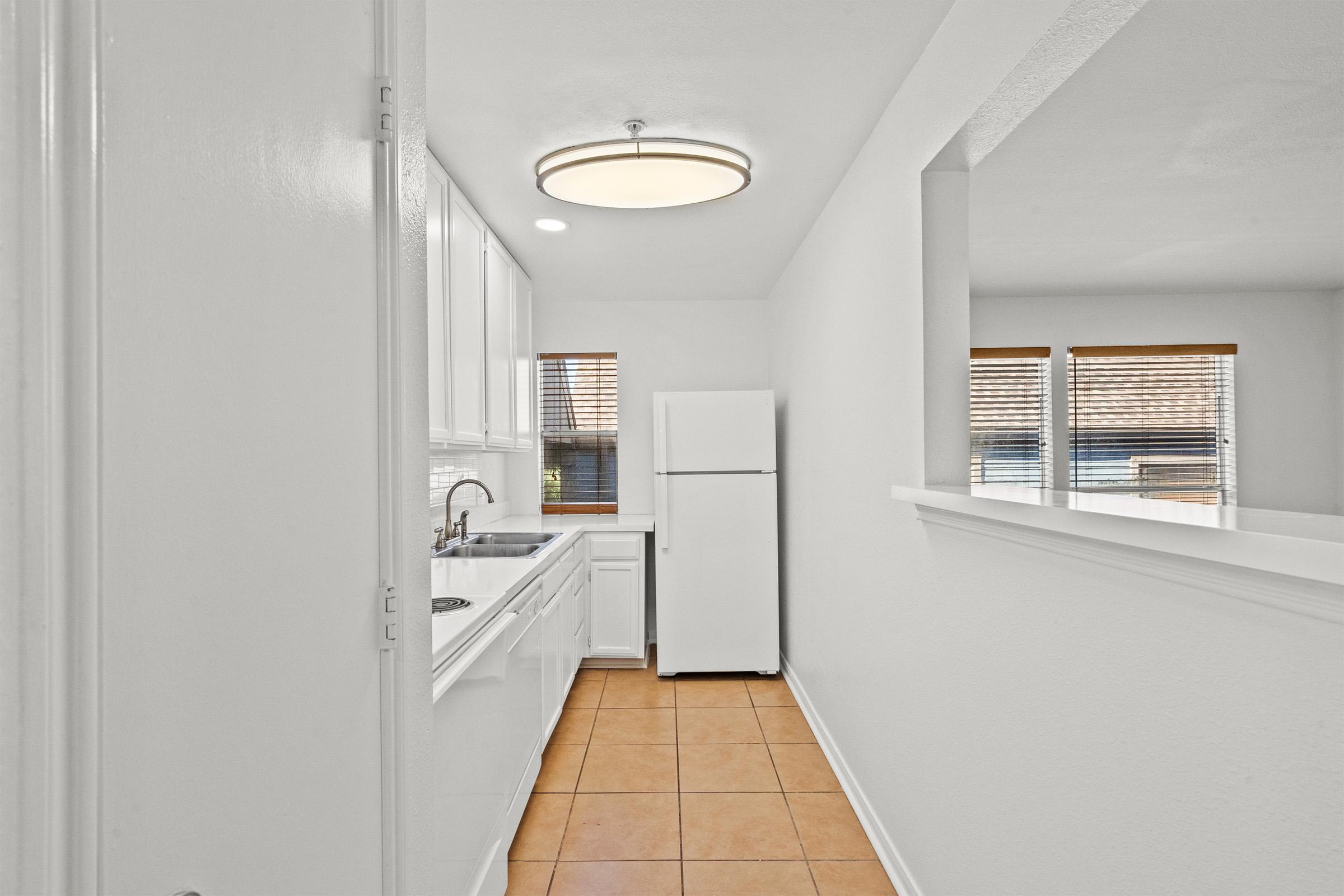 A bright, modern kitchen featuring white cabinetry, a stainless steel sink, and a white refrigerator. The tiled floor and large windows provide natural light, while a contemporary ceiling light fixture enhances the ambiance. The overall aesthetic is clean and inviting.