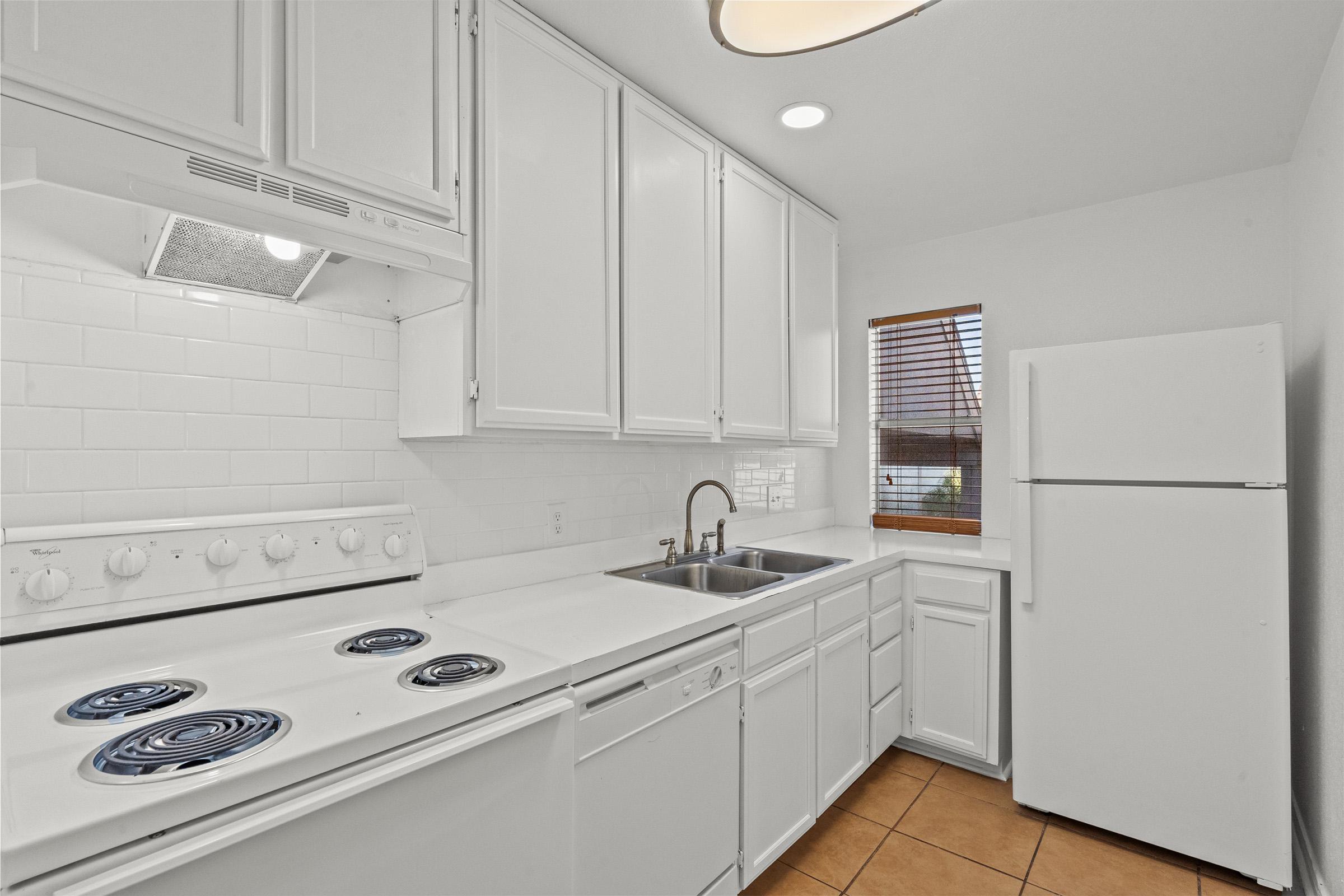Bright and modern kitchen featuring white cabinetry, a white refrigerator, a sink with a faucet, and a stove with four burners. A window with blinds allows natural light to enter, and the floor is tiled in a warm, brown color. The overall aesthetic is clean and minimalist.