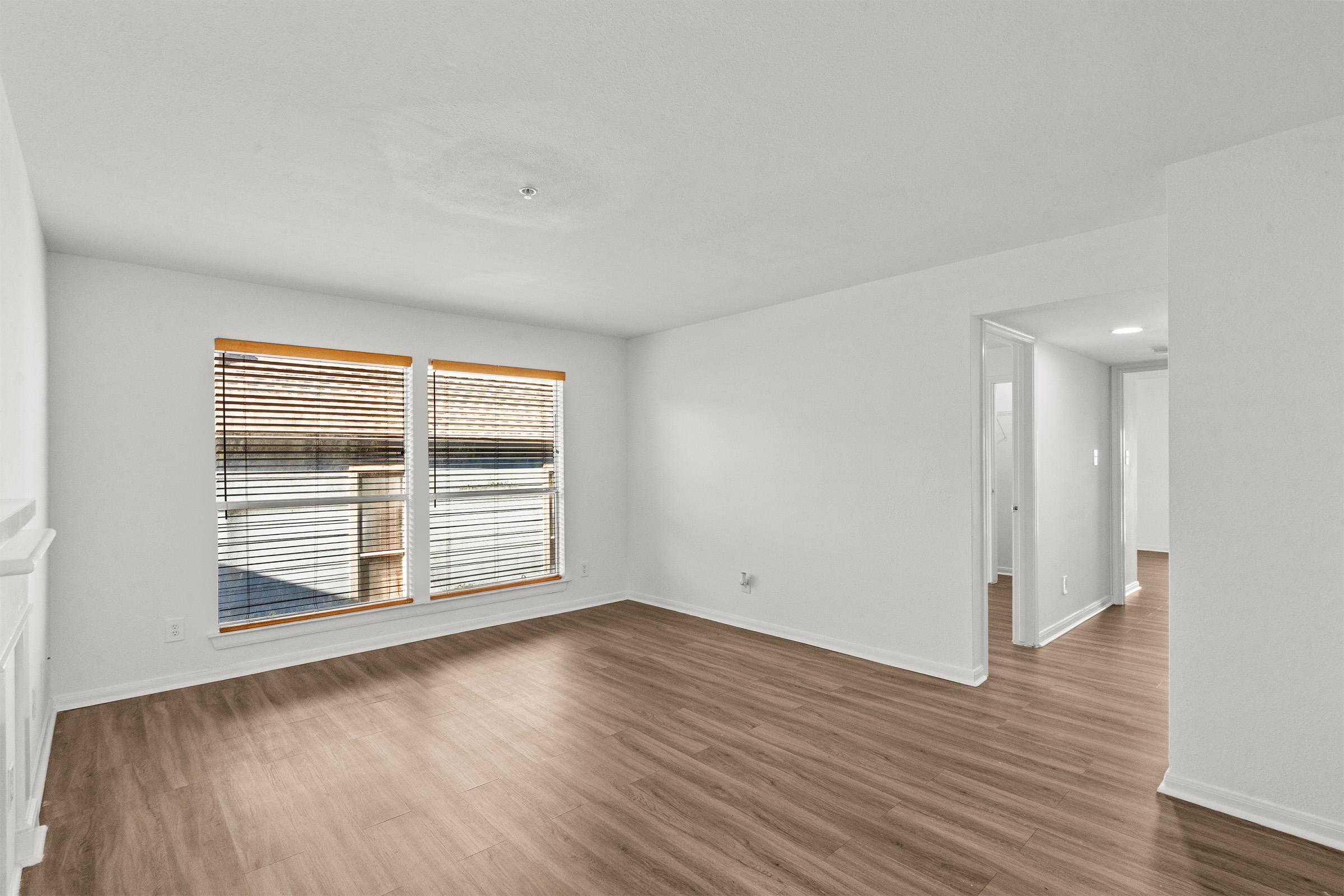 A bright, empty room with light-colored walls and wooden flooring. Two windows with white blinds allow natural light to enter. The space features a plain white wall and an open area leading to another room, creating a spacious and inviting atmosphere.