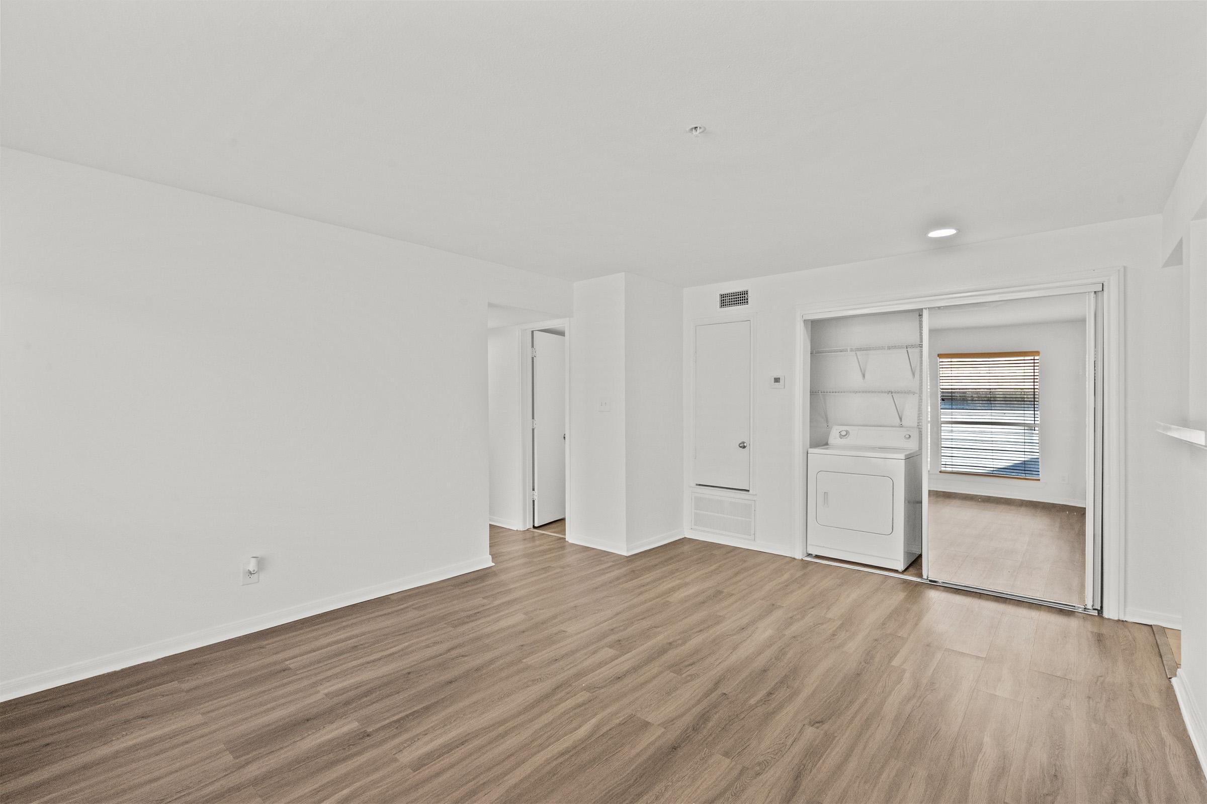A bright, empty room with light-colored walls and wooden flooring. A doorway leads to another space featuring a washing machine. Natural light streams through a window, illuminating the area. The overall atmosphere is clean and modern, with minimal furnishings.