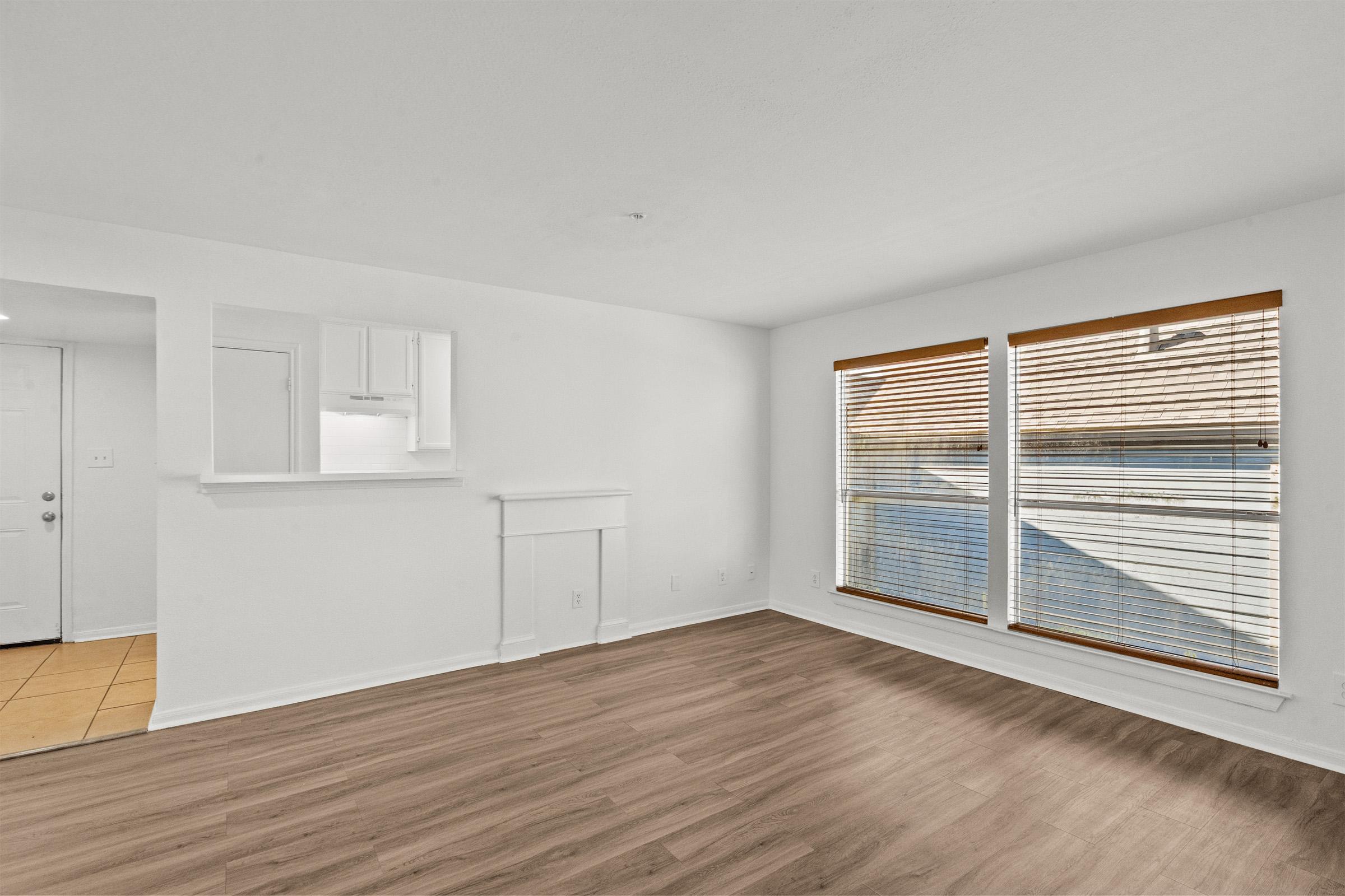 A spacious, well-lit living area featuring two large windows with wooden blinds, a light-colored wall, and a wood floor. To the left, there's a small open area leading to a kitchenette with a bar-style counter. The space has a clean and modern feel with minimal furnishings.