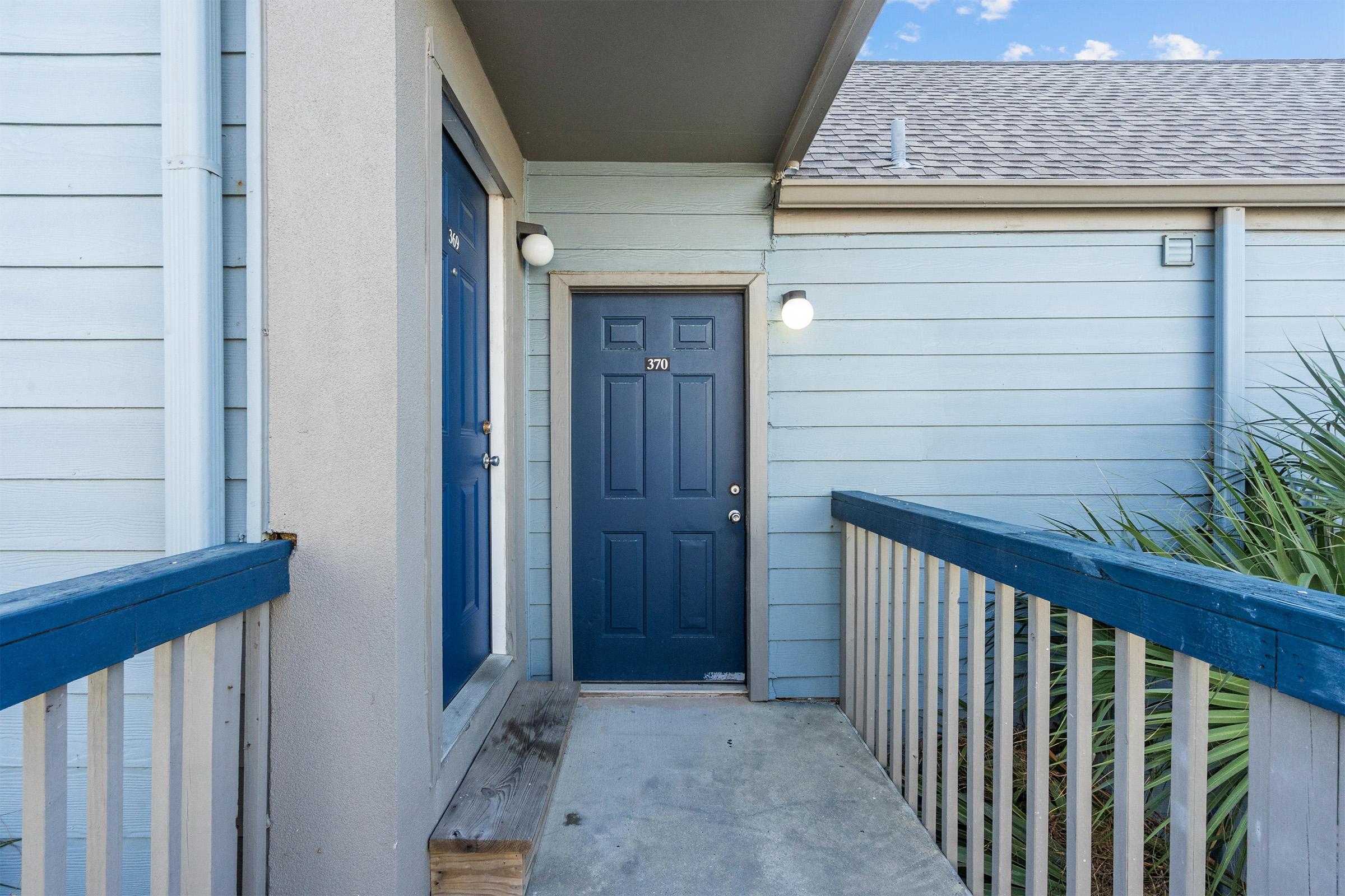 A blue door is framed by light blue siding and is accessed via a wooden ramp. The exterior features simple design elements with a light fixture on either side of the door and greenery visible nearby. The scene appears bright and inviting.