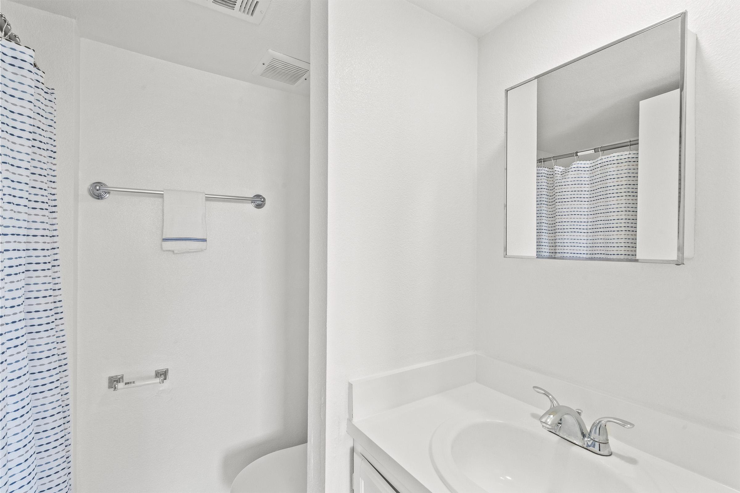 A small, clean bathroom featuring a white countertop with a faucet, a mirror above it, and a shower area with a blue and white striped curtain. The walls and surfaces are primarily white, creating a bright and minimalist atmosphere.